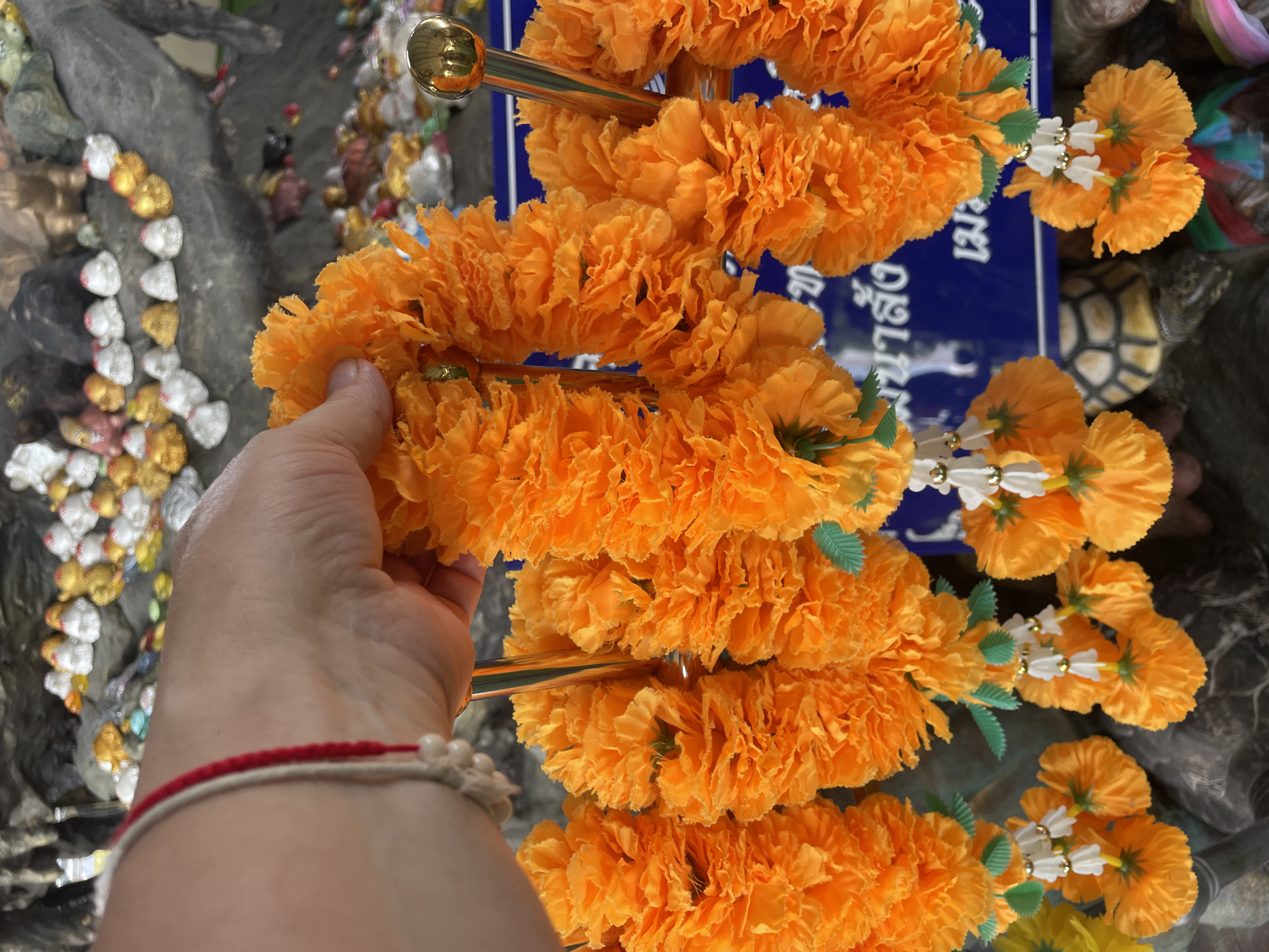 A close-up shot shows a person's hand holding an orange marigold garland. The garland is made of many marigold flowers strung together, with decorative white and gold beads interspersed. The background reveals a stone structure adorned with numerous small, colorful figurines, some resembling turtles and others more abstract. A blue sign with Thai script is visible behind the garlands. The overall scene suggests a cultural or religious setting, possibly a shrine or temple in Tambon Na Chom Thian, Thailand, given the context of marigold garlands being used in such places.