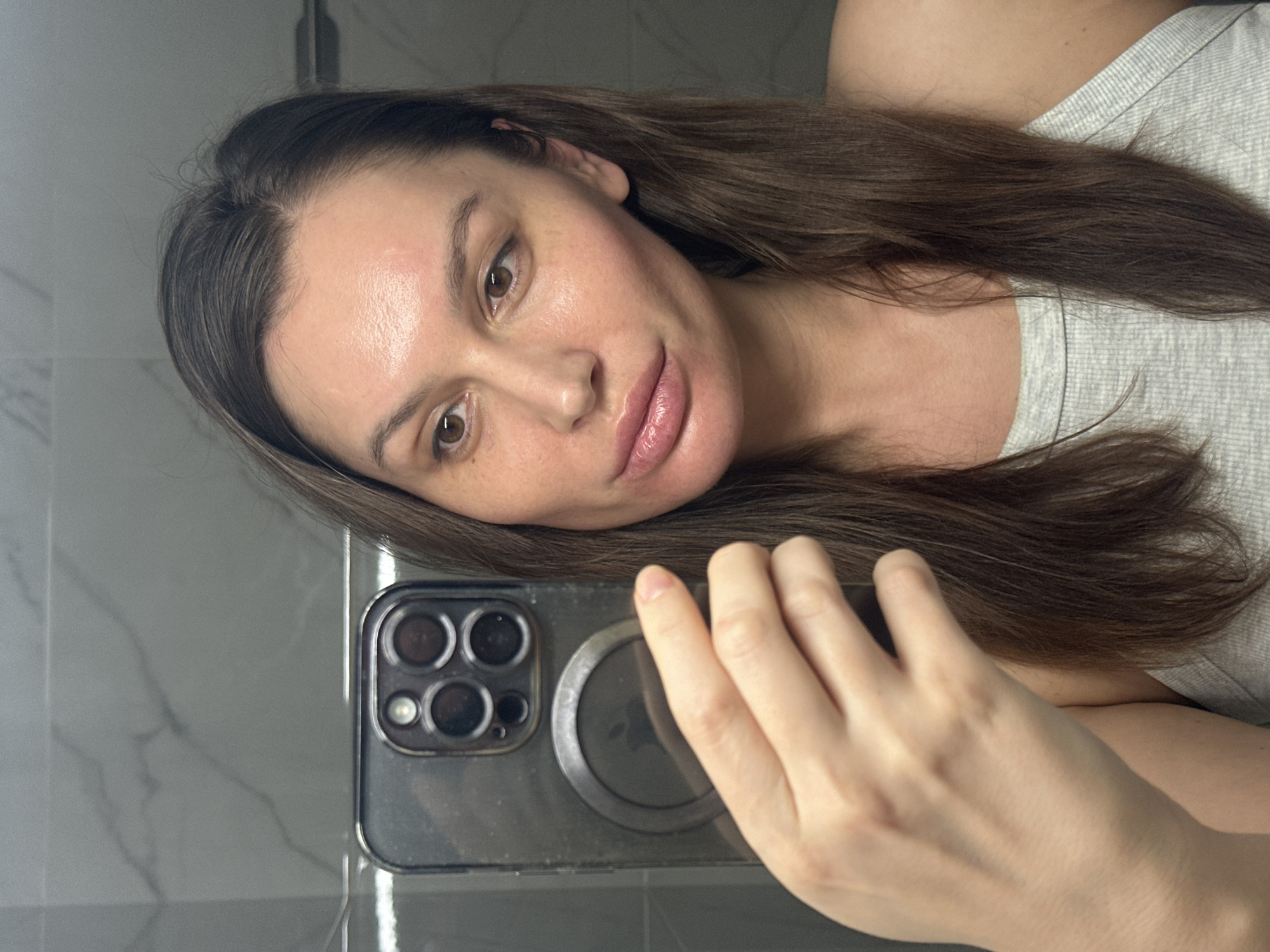 An indoor mirror selfie features a woman with long, dark brown hair and a neutral to slightly pensive expression, captured in Moscow, Russia. She looks directly at the camera, her head tilted to her right. Her lips are slightly parted, revealing a glossy finish, and her brown eyes are prominent. Her skin shows a natural sheen on her forehead and cheeks, with a subtle patch of redness above her left eyebrow. She is dressed in a light gray, ribbed or textured top.
Her right hand holds a dark-cased smartphone, partially visible in the lower right foreground. The phone's back clearly displays a multi-lens camera array, including three larger lenses and a smaller light or sensor, alongside a prominent circular magnetic attachment (likely MagSafe). The phone's reflective surface shows distortions of the surrounding environment.
The background consists of a smooth, grey surface with subtle white marbling patterns, indicative of a modern bathroom or vanity setting. The lighting is soft and even, highlighting her face and the reflective phone. No text is visible. The scene conveys a calm and candid moment.