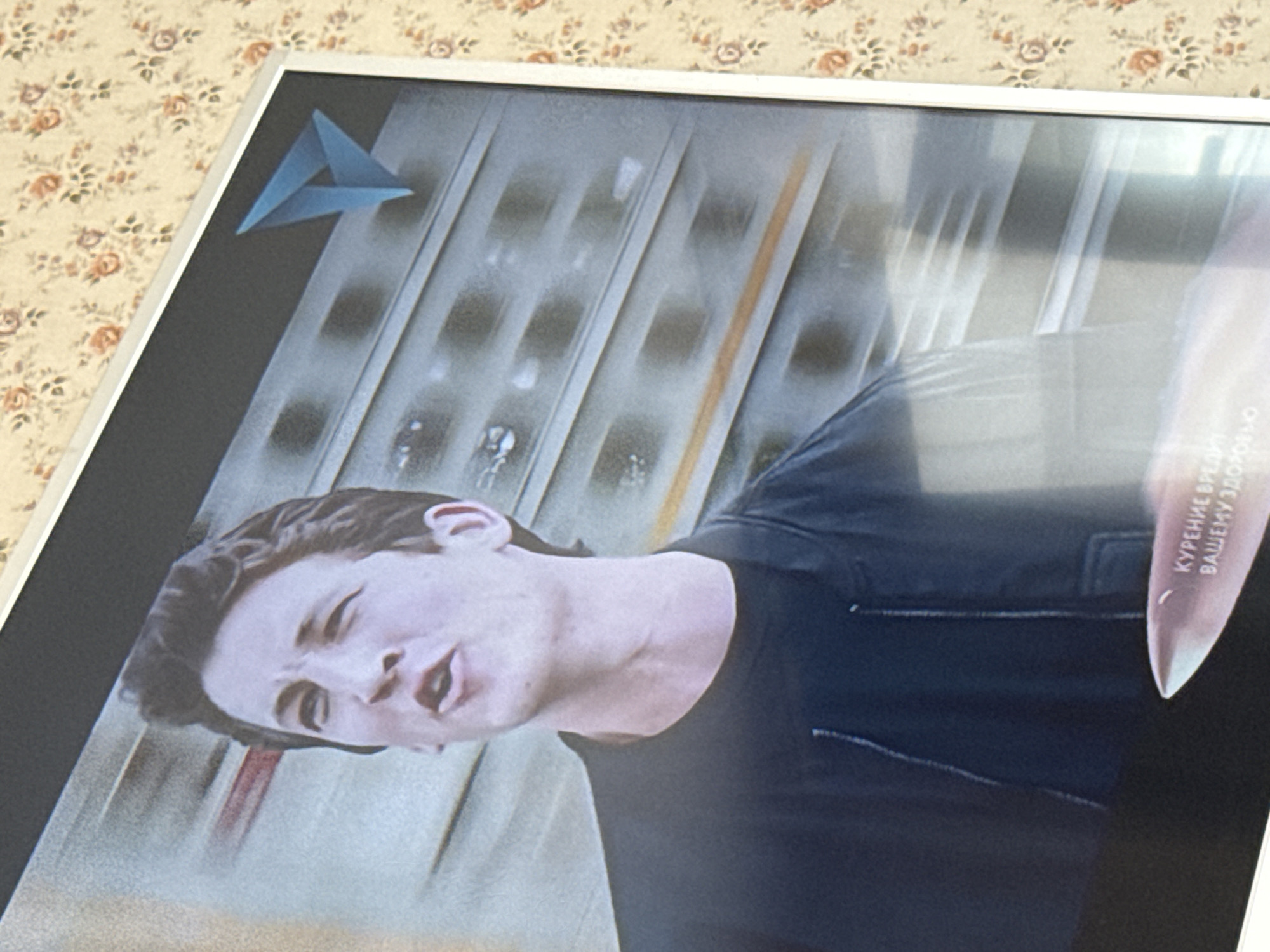 A close-up shot of a television screen displays a man in a dark shirt. Behind him, there's a blurry background of vertical and horizontal lines, potentially suggesting a train or subway setting. The upper left corner of the screen shows a blue triangle logo. The lower right corner of the screen shows the warning: "КУРЕНИЕ ВРЕДИТ ВАШЕМУ ЗДОРОВЬЮ" (Smoking is harmful to your health). The television is set against a backdrop of floral-patterned wallpaper.
Location: The text on the screen is in Russian, but Pushkino is not discernible in the image.
