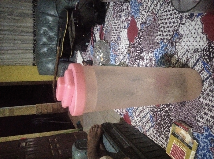 This indoor photograph captures a casual scene within what appears to be a residence in Kalirejo, Indonesia. The dominant visual element is a large, translucent cylindrical bottle with a distinctive pink, multi-tiered lid, possibly a shaker or water bottle, lying horizontally across the midground. It rests upon a vibrantly patterned textile, strongly resembling traditional Indonesian batik, featuring intricate designs in shades of blue, red, white, and black, which serves as a significant location cue.

In the background, a dark, possibly leather or faux-leather armchair or sofa is visible to the left, with a black acoustic guitar leaning against it. A bright yellow wall provides a contrasting backdrop further to the left. Towards the center-right, a shiny, textured object, possibly an ashtray or decorative item, is partially visible.

In the bottom right foreground, a pair of bare feet are seen, implying the presence of a person relaxing. Adjacent to the feet, a cigarette pack is clearly visible, with the prominent text "DJI SAM SOE," a popular Indonesian brand. The overall lighting suggests an indoor setting, not brightly illuminated, contributing to a relaxed and personal atmosphere. No specific activity beyond casual relaxation is depicted, but the collection of personal items like the guitar, bottle, and cigarettes hints at leisure.