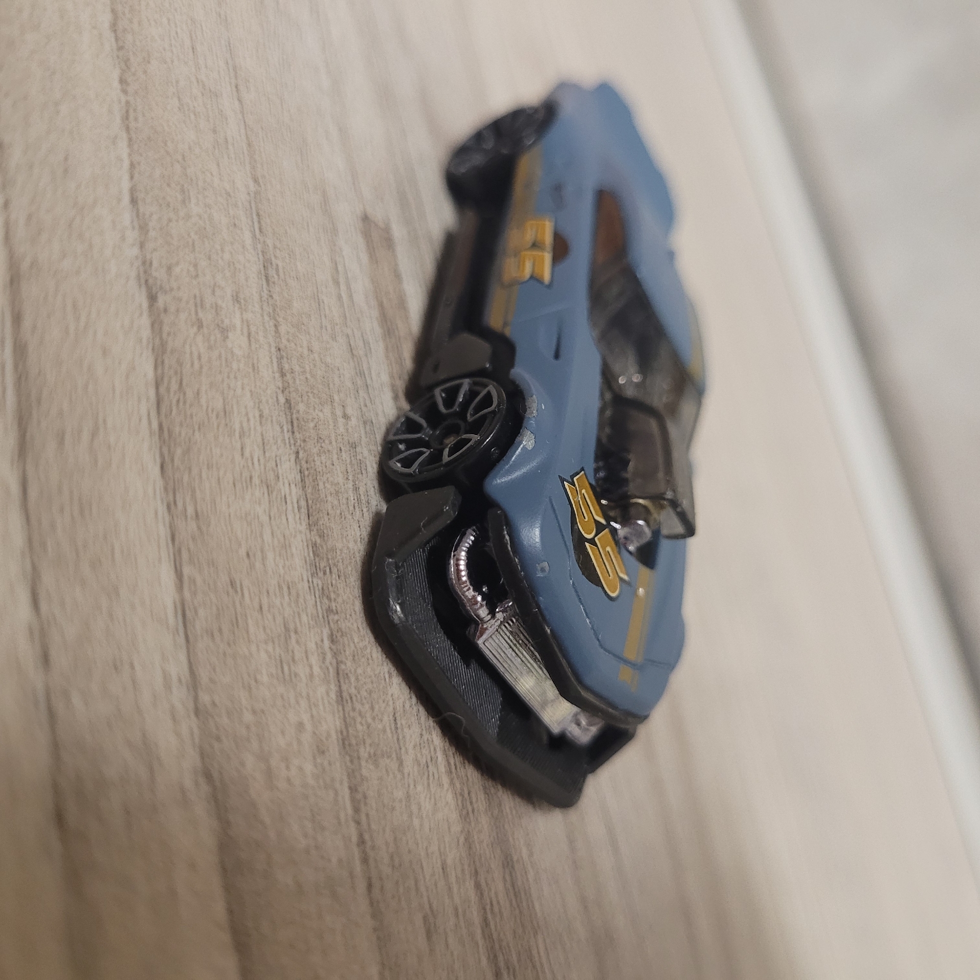 A blue toy race car with yellow accents is photographed at an angle, lying on its side. The car is positioned on a surface that appears to be a light-colored wooden floor with visible grain. The toy car has detailed wheels and what looks like an engine or exhaust pipe visible underneath. There is yellow text with the number "55" on the side of the car. The lighting suggests an indoor setting, possibly artificial light. The background is blurred, but appears to be a lighter surface, possibly a wall. The image is focused on the toy car, highlighting its details.
