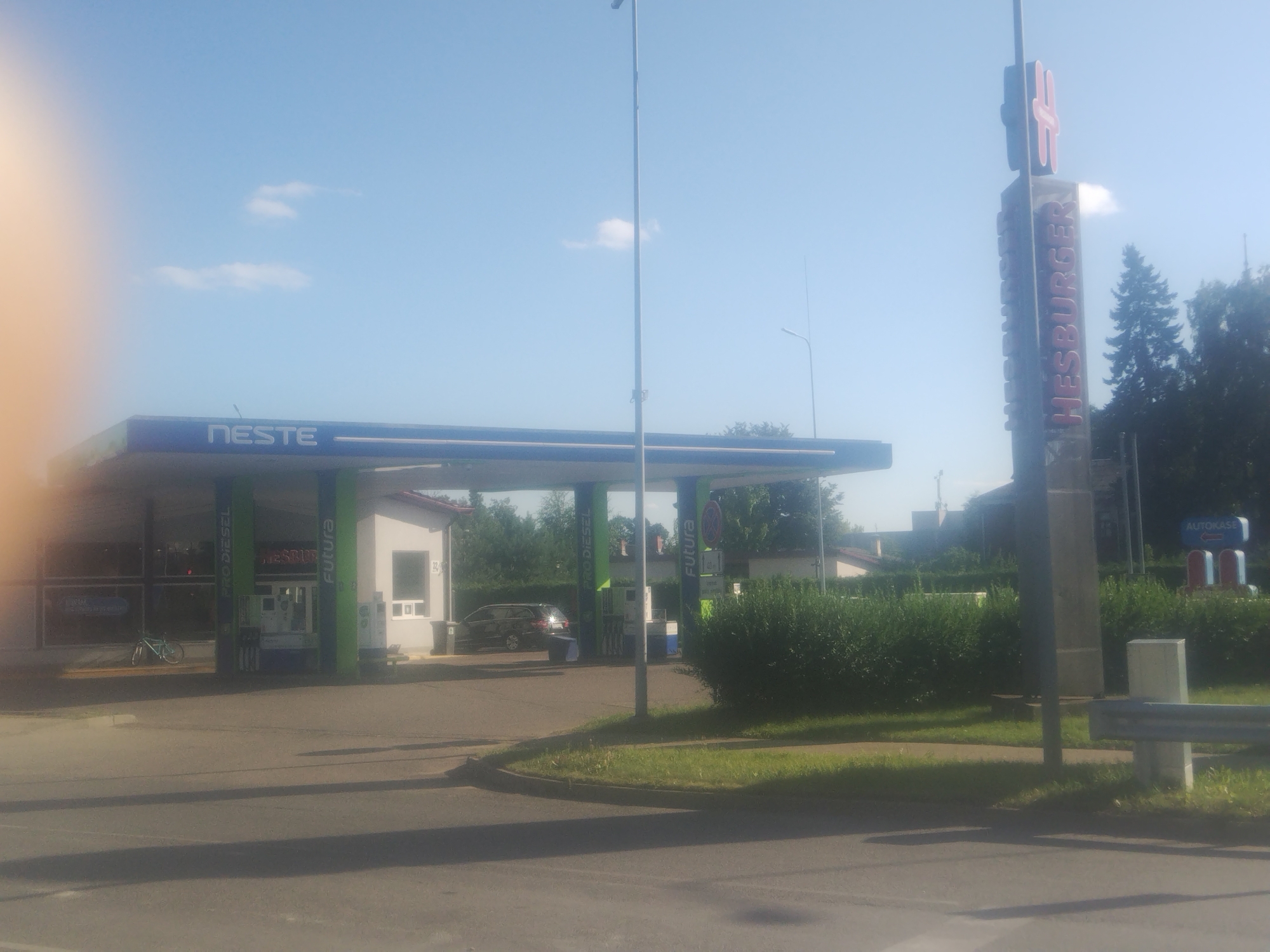 The image depicts a Neste gas station and a Hesburger fast-food restaurant, likely located in Daugavpils, Latvia, on a sunny day. The Neste station has a blue and green canopy with the "NESTE" logo visible. Several green pillars support the canopy, with some displaying text like "PRO DIESEL" and "FUTURA". A white attendant booth is situated near the pumps. In front of the station, a dark-colored car is parked, and a bicycle is leaning against a wall to the left. To the right of the gas station, a prominent sign for Hesburger, with its characteristic red "H" logo and red lettering spelling "HESBURGER", stands tall. The surrounding environment includes a well-maintained green hedge, grass, and trees in the background. A street or road is visible in the foreground, casting shadows. The sky is bright blue with a few scattered clouds. An "AUTOKASE" sign with arrows is also visible in the distance.