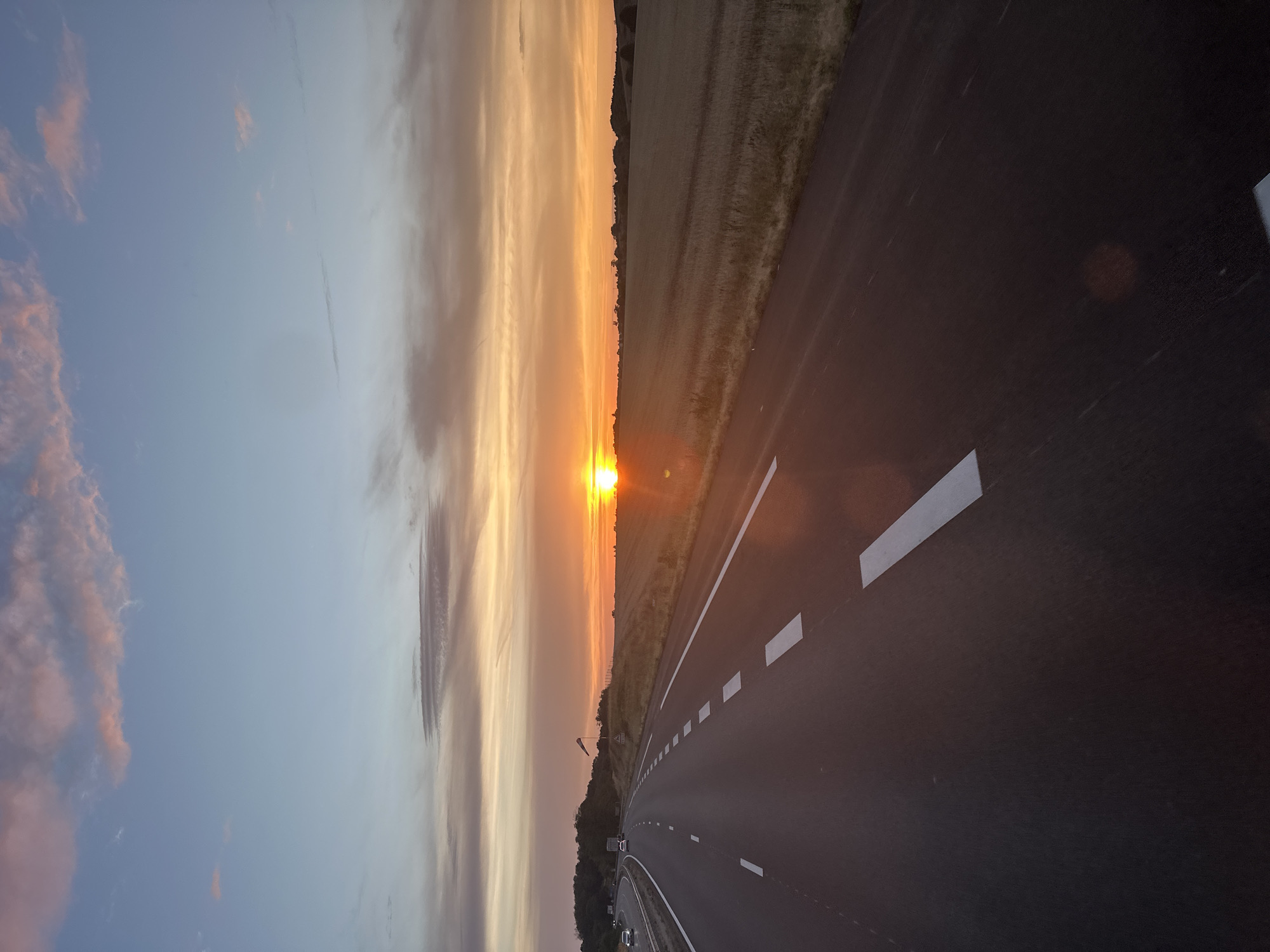A vertical, eye-level, full shot captures a road and a sunset over a field in Authume, France. The paved road occupies the left foreground, curving to the left as it recedes into the distance. White dashed lines mark the lanes. On the right side of the road is a field of dry grass, stretching to the horizon. The sky is filled with a vibrant sunset, transitioning from orange at the horizon to pale blue at the top of the frame. Wispy clouds are scattered across the sky, tinged with pink and orange. A few trees and a small hill are visible in the distance on the left side of the road, and a single vehicle is driving on the highway. A flag on a pole stands on the right side of the road. The image appears to have been taken during golden hour, with the sun low in the sky.