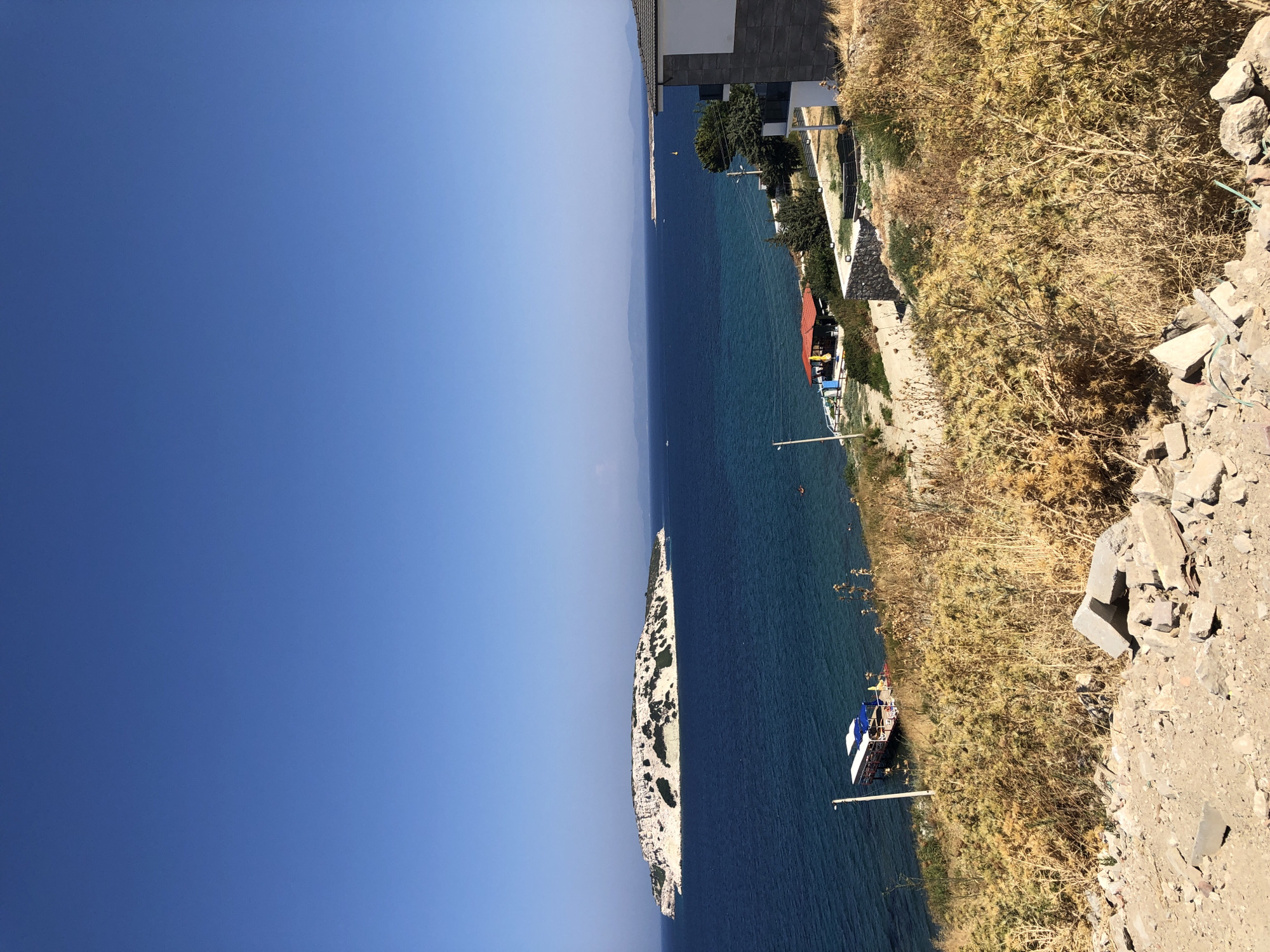An eye-level, wide-angle shot captures a scenic coastal view under a clear blue sky. In the foreground, dry, golden brush and scattered rocks cover the ground, suggesting a rugged, natural landscape. A paved path winds down the slope.
Further down, a body of deep blue water stretches towards the horizon. An island, dotted with white buildings and patches of greenery, sits in the middle ground of the sea. A small boat with a white roof and blue accents is docked near the shore.
Along the coastline, a small beach area is visible with a structure featuring a red roof and yellow umbrellas, possibly a beachside cafe or bar. To the right, a modern building with grey stone accents and a white facade is situated on a higher elevation. The distant horizon shows a hazy outline of landmasses under the bright sun. The location is identified as Karaburun, Turkey.
