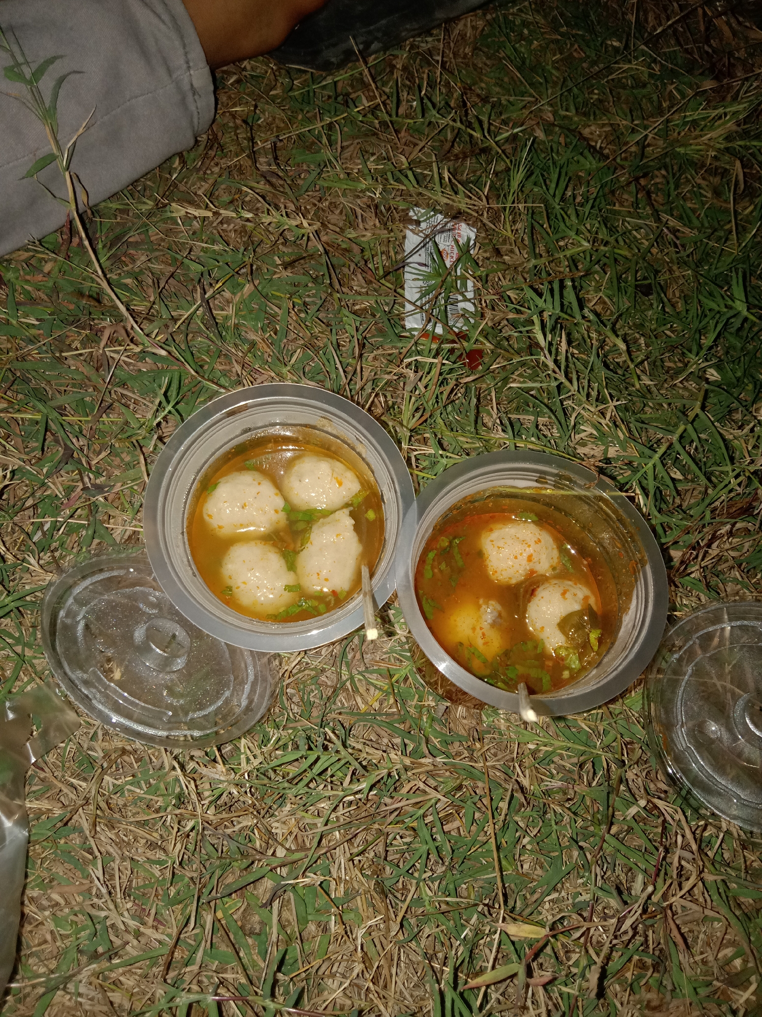 Two plastic cups filled with meatballs and broth are sitting on the ground amongst dry and green grass. One cup, on the left, contains four meatballs and is partially obscured by a clear plastic lid placed beside it. The other cup, on the right, contains three meatballs and appears to have a spoon sticking out of it. A person's leg, clad in grey pants, is visible in the upper left portion of the frame. The scene appears to be outdoors in a grassy area.