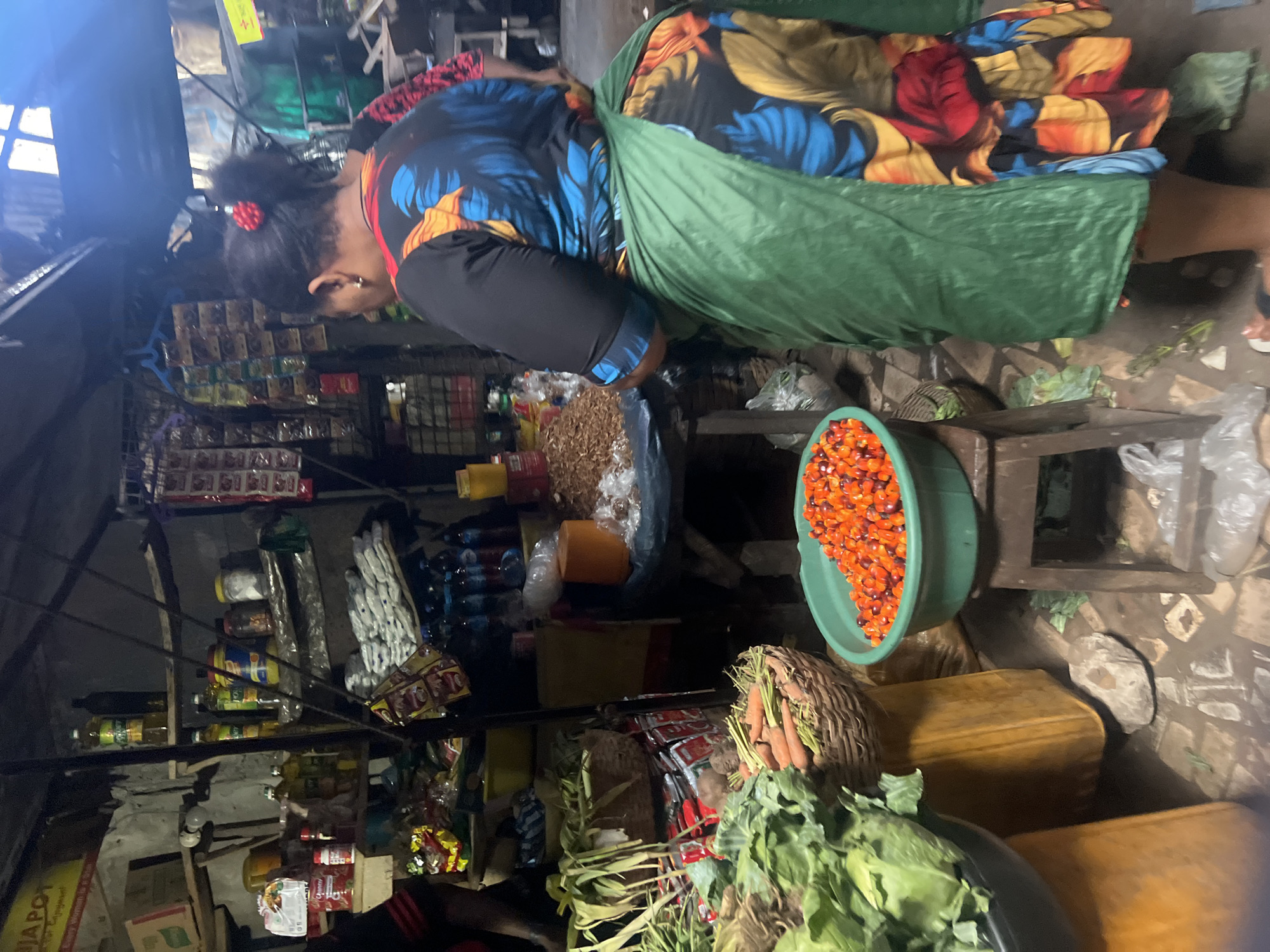 A woman wearing a floral patterned top and a green apron leans over a table laden with goods inside a market stall. The stall displays a variety of items, including bottled goods, dried goods, palm fruit in a green bucket, and leafy vegetables. The market stall is located in Nkpor, Nigeria. The scene appears to be a typical market setting, with limited space and a focus on commerce.