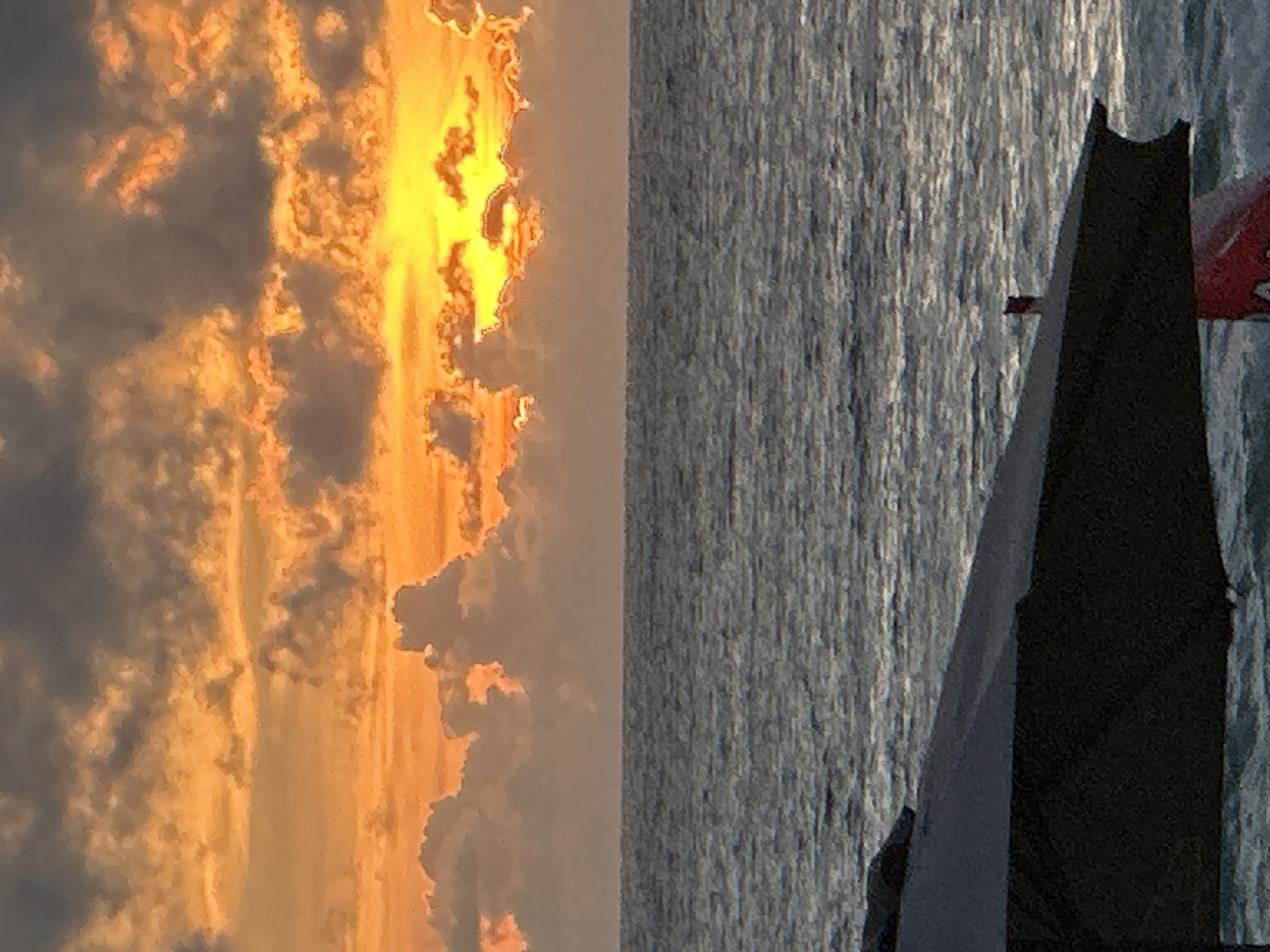 This image shows a dramatic sunset over a body of water, possibly the Andaman Sea, near Karon, Thailand. The left side of the frame is dominated by a vibrant sky filled with clouds tinged with fiery orange and golden yellow, suggesting the sun is low on the horizon. These warm hues contrast with darker, grey clouds, creating a dynamic and textured sky.
The right side of the image features a textured surface of water, appearing somewhat choppy and reflecting the light in vertical streaks, indicative of a moderate breeze. Partially obscuring the water and sky is a dark sail or hull of a boat, possibly a sailboat due to the triangular shape and the presence of a red flag with white markings, which might be a national flag or a team pennant. The boat appears to be moving or anchored in the water.
The overall scene evokes a sense of peacefulness and awe, characteristic of a beautiful sunset. The light suggests it is late afternoon or early evening. No people are clearly visible. The red and white flag is the only potentially legible element, but its details are not clear enough to identify specific text or symbols.