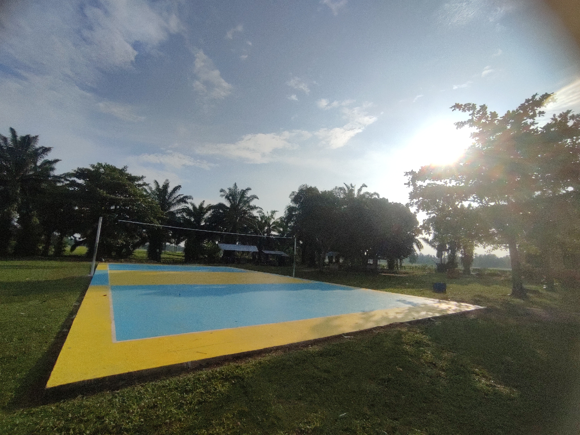 This wide-angle outdoor shot captures a vibrant volleyball court set amidst a verdant landscape in Blangriek, Indonesia. The court features a distinct design with light blue and yellow painted sections, delineated by white lines, and is equipped with a net and supporting posts. No people are visible on the court or in the surrounding area, suggesting a moment of quiet before or after activity.
The court is surrounded by lush green grass and numerous trees, including prominent palm trees, which frame the scene and extend into the background. Hints of distant structures or open fields can be seen beyond the tree line.
The image is illuminated by bright, direct sunlight, suggesting either morning or late afternoon. The sun, positioned towards the upper right, creates a notable lens flare, adding a warm glow to that side of the frame. The sky is a clear light blue with scattered white clouds. The overall scene conveys a sense of natural beauty and readiness for recreational use. No text is visible in the media file.