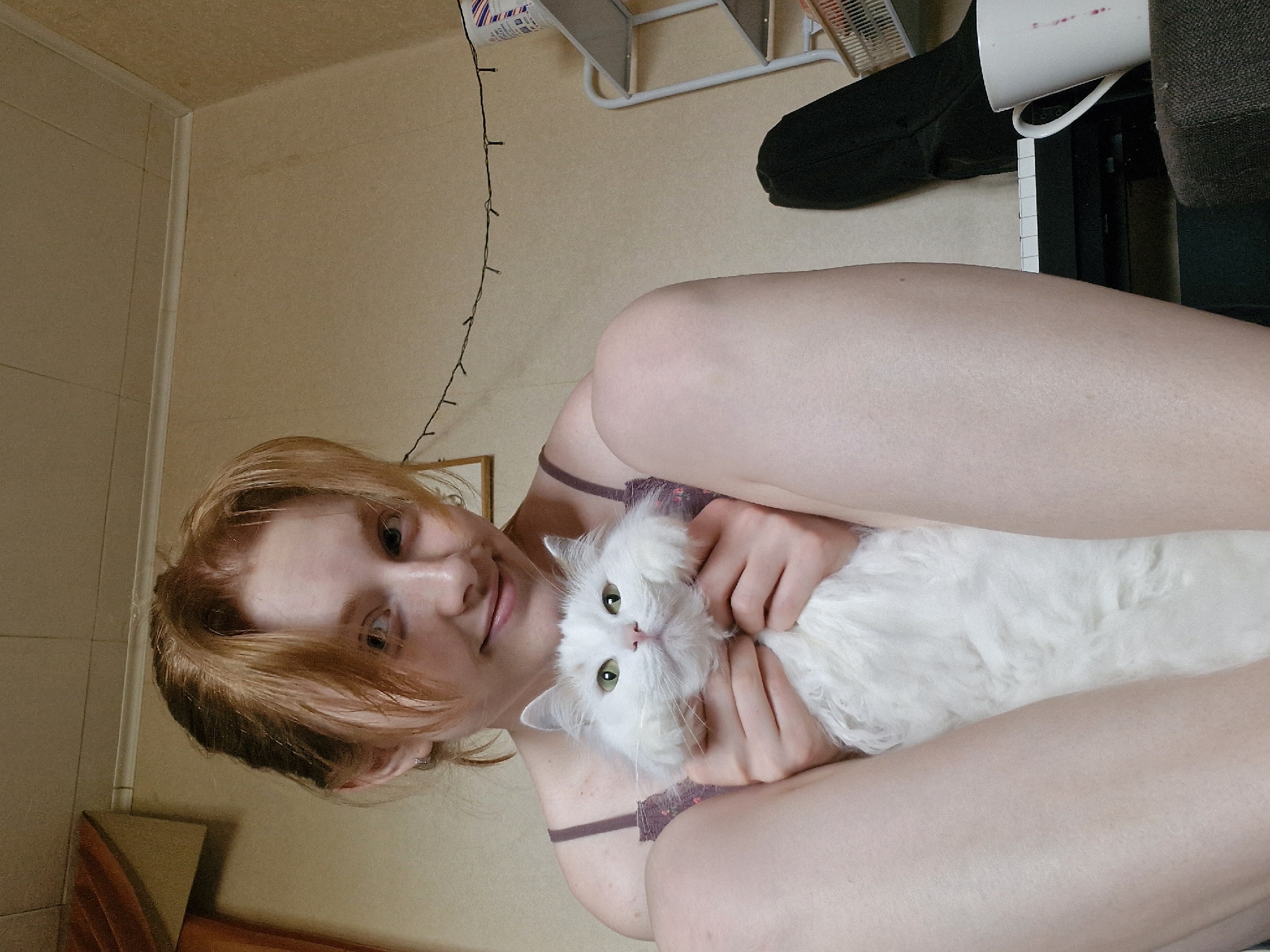 A woman is holding a white, fluffy cat. The woman has fair skin and reddish-blonde hair. She is wearing a camisole. The environment appears to be an indoor residential space with beige walls. The location is unspecified, and there's no information to definitively place it in Yekaterinburg, Russia. Visible in the background are a metal shelf unit, a black object (possibly clothing), a white mug, and a portion of a keyboard.