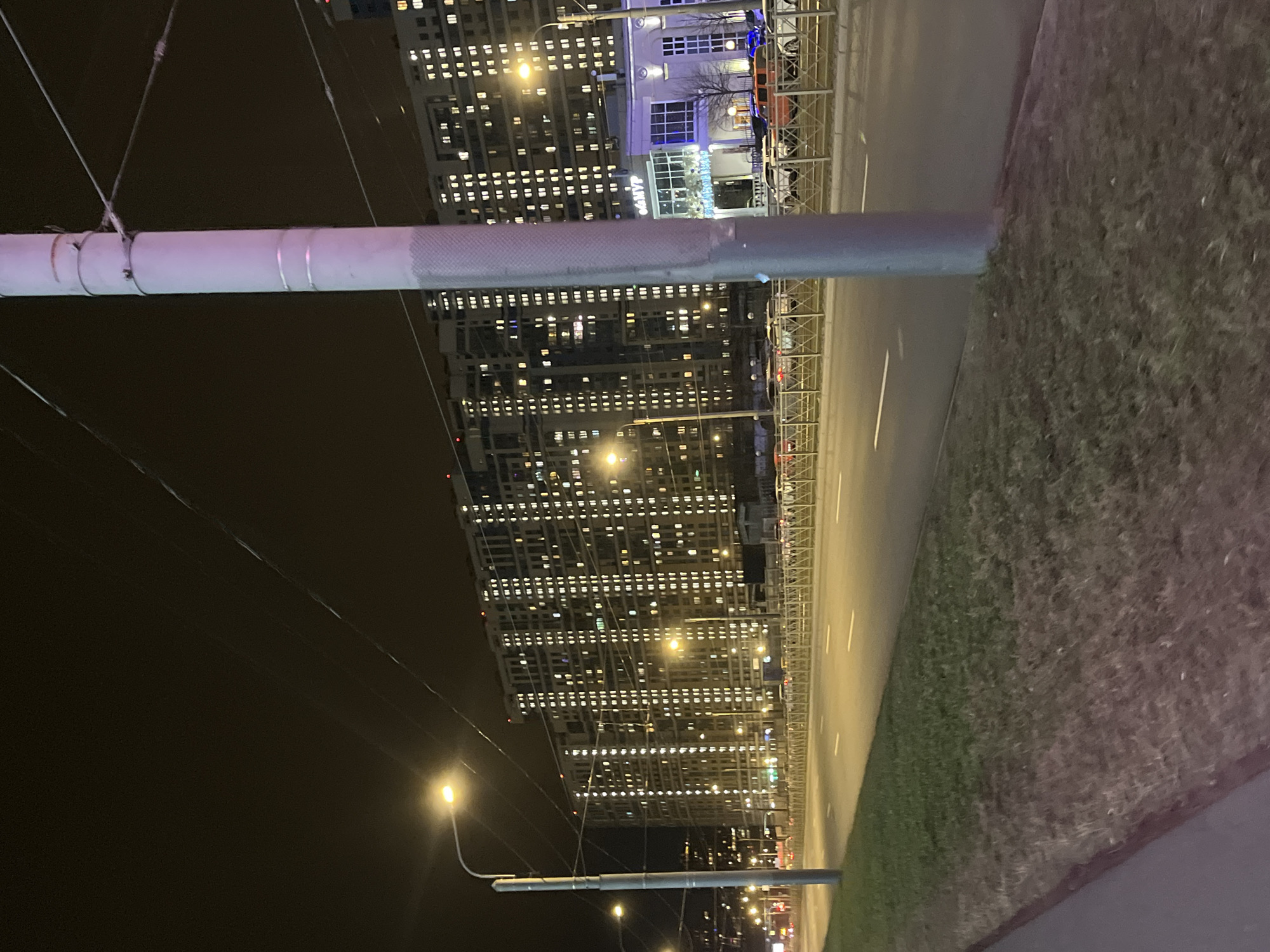 The image, taken at night, shows a view in Saint Petersburg, Russia. In the foreground, a metal structure or post dominates the scene, with cables extending from it. A road with white lines runs along the right side of the image, bordering a patch of vegetation. Across the road, tall buildings with numerous illuminated windows create a cityscape backdrop. Streetlights are visible along the road, and a lit sign that reads "O&MyP" is present on one of the buildings. The sky is dark.
