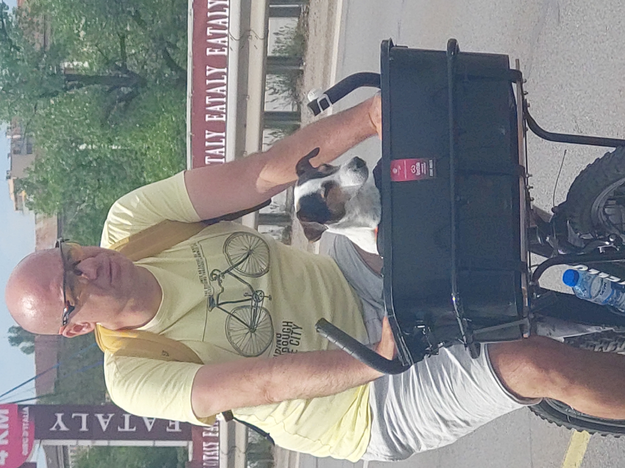 A medium shot captures a bald man in a light yellow t-shirt and grey shorts on a bicycle with a black cargo basket. A white and brown dog sits inside the basket. Behind the man is a sign for "EATALY". Another sign indicates the location is 4 KM from "Eataly Sofia, Bulgaria". The background includes green foliage and a light blue sky. The man appears to be riding the bicycle.