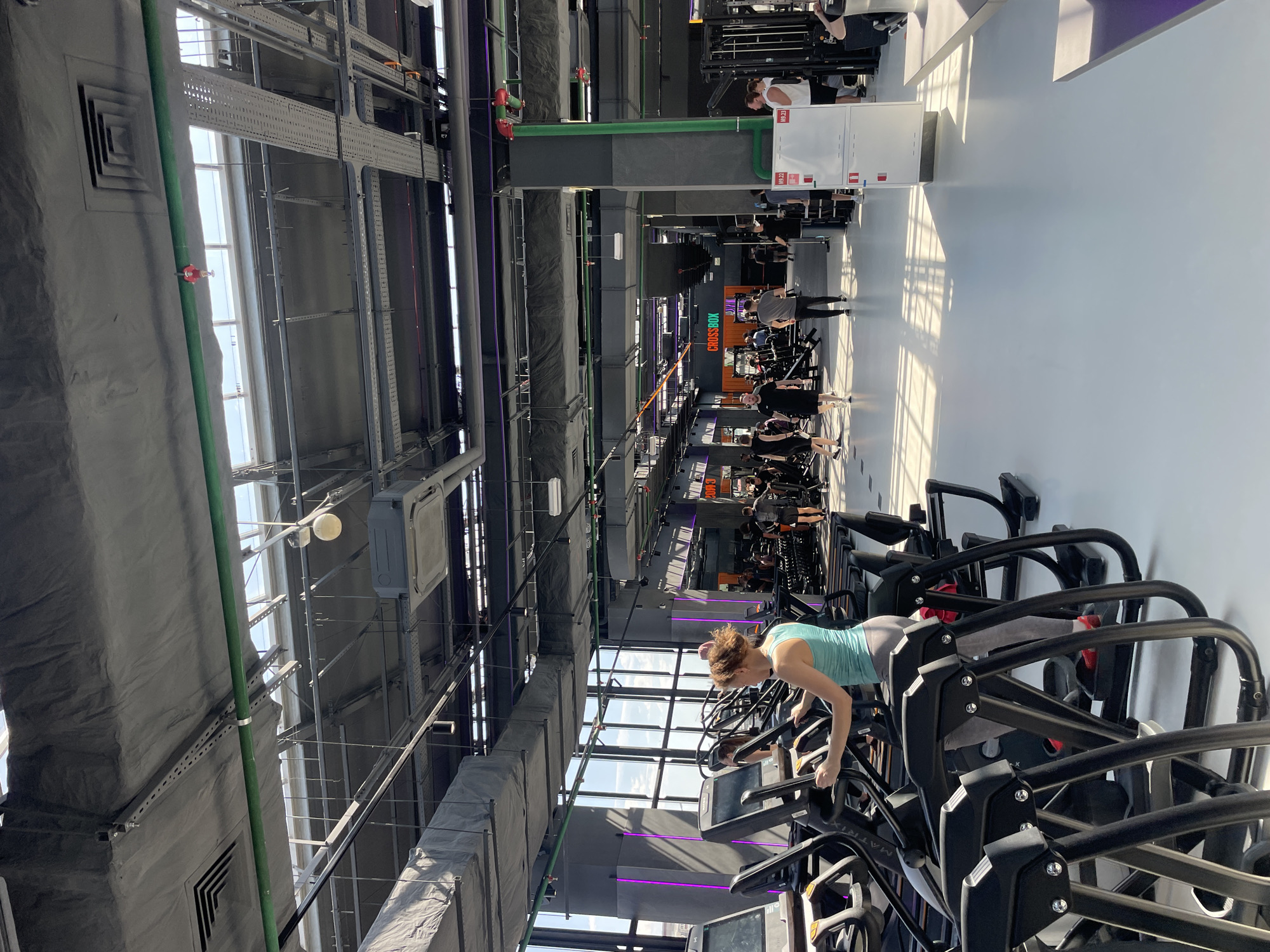 An interior shot depicts a gym, likely in Kazan, Russia, given the Cyrillic script partially visible on a sign. A woman in a blue top and grey leggings is on a stair-climbing machine. The gym features high ceilings with exposed ductwork and ventilation systems. Several other individuals are exercising in the background, operating various weight training machines. Natural light streams in through the windows. The floor is a light grey color. The gym is modern and well-equipped.