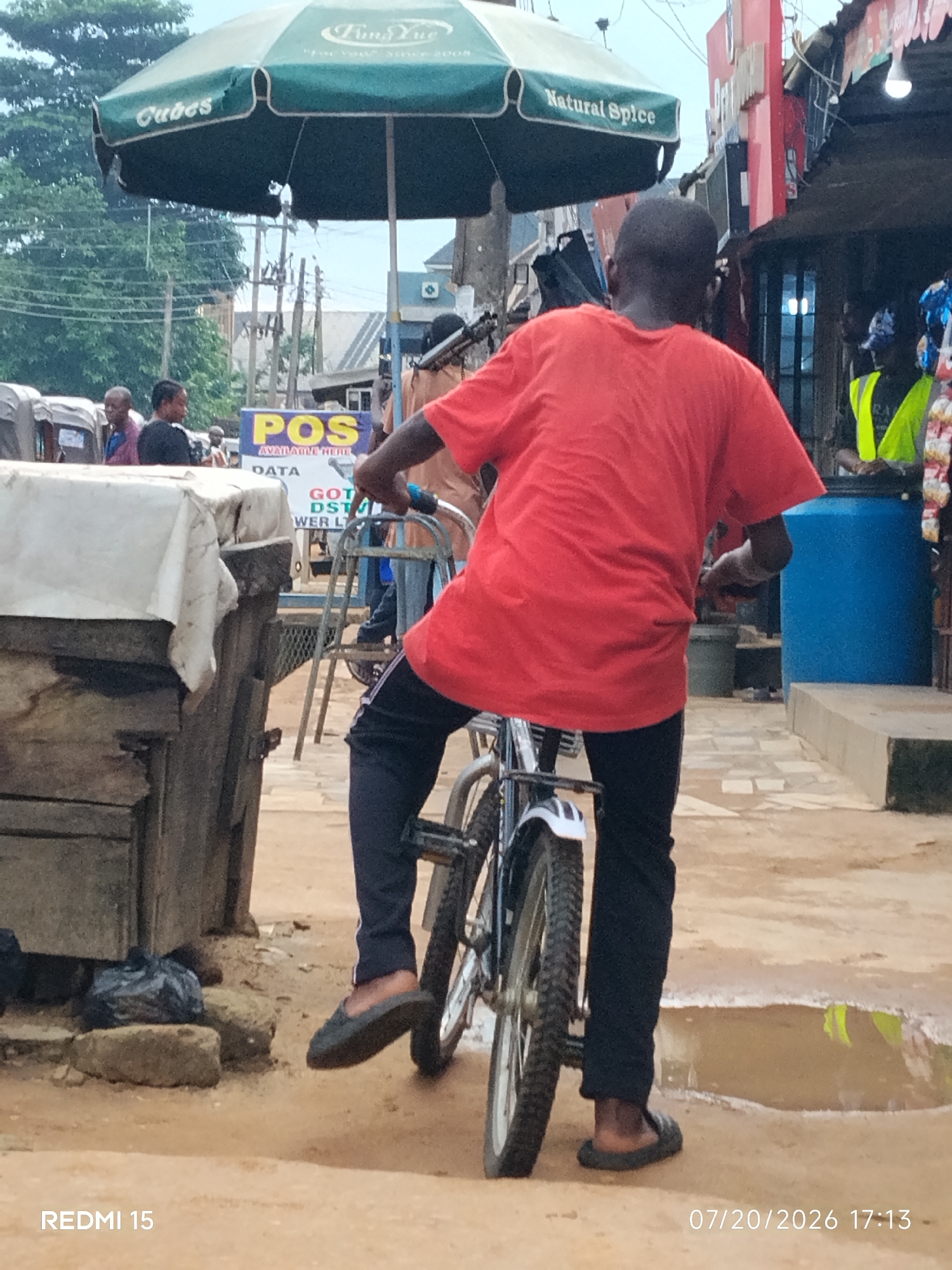 In Umuode, Nigeria, a young Black male is depicted from behind, wearing a bright orange t-shirt and dark pants, riding a bicycle. He is in motion, with his right foot on the pedal and his left foot slightly extended. The bicycle has a silver frame and dark tires. The ground beneath him is a mix of dirt and puddles, suggesting recent rain. To his left, a wooden structure covered with a white cloth stands. In the background, a large green umbrella with the words "Cubes" and "Natural Spice" printed on it is visible. A blue barrel is situated to the right of the frame. Other individuals and market stalls are also present in the background, hinting at a bustling marketplace environment. The image is dated 07/20/2026.