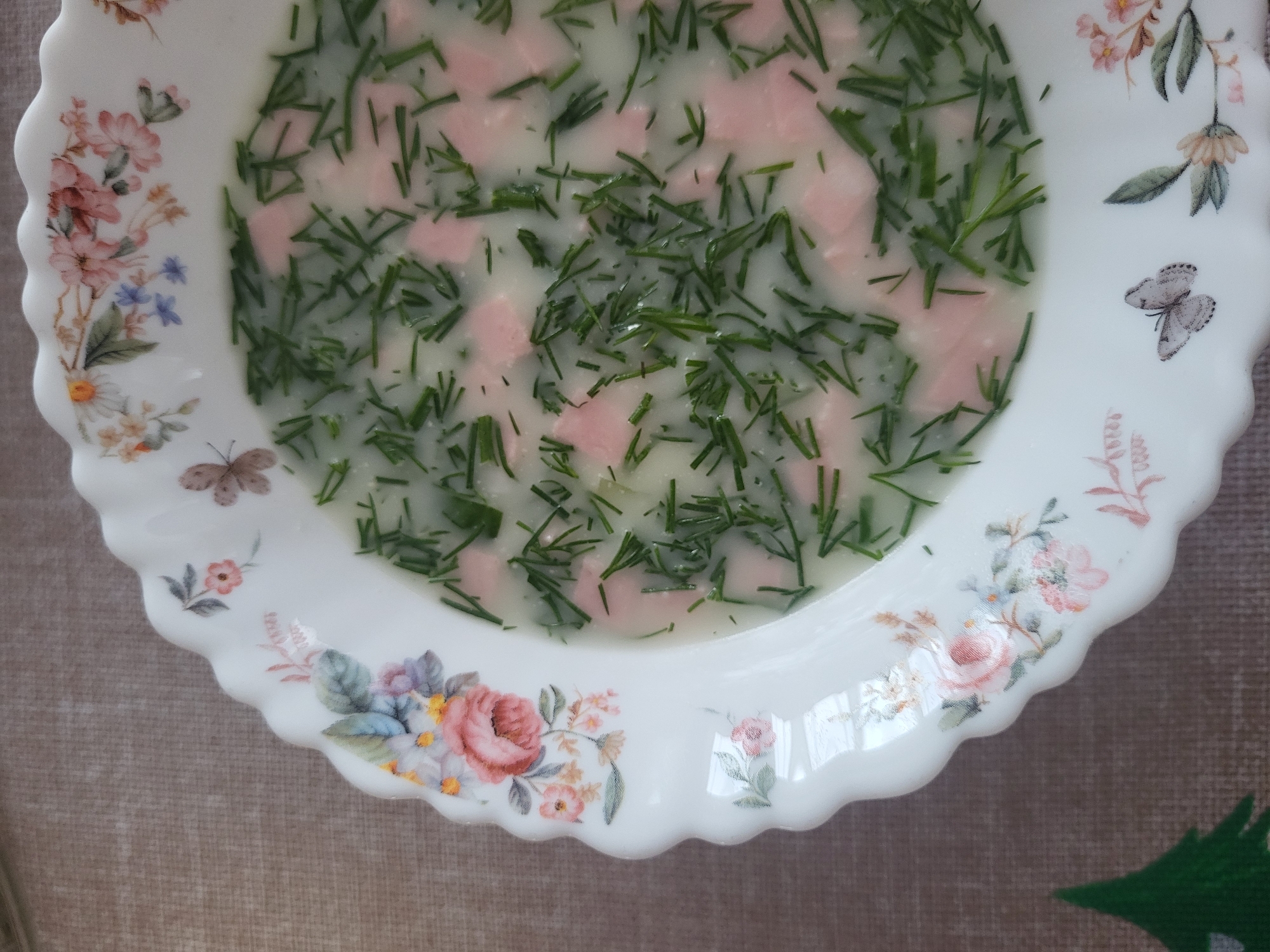 A close-up shot shows a bowl of cold soup, potentially Okroshka, on a floral patterned plate, placed on a beige textured surface. The soup contains chopped dill and diced ham or sausage. The plate's rim is decorated with painted flowers including roses and daisies, as well as a butterfly. The location, Зипуново, Russia, is likely the origin of the image.