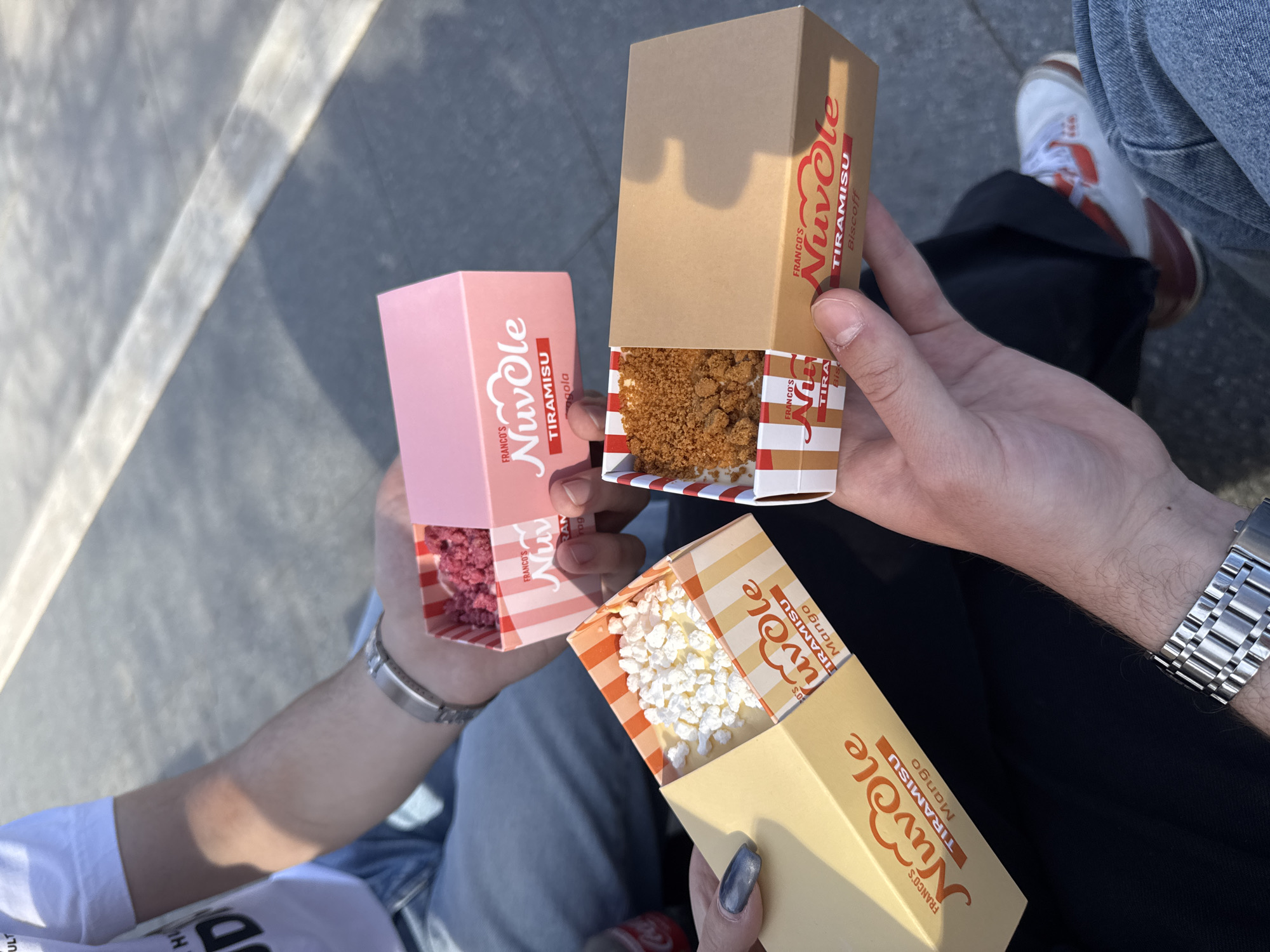 Three people are holding small boxes of Franco's Nuvole Tiramisu in different flavors: biscof, fragola, and mango. The people are standing in what appears to be an urban environment, likely in Sofia, Bulgaria, judging by the text on the boxes. The person holding the mango-flavored tiramisu is wearing a metal watch. The person holding the fragola-flavored tiramisu is wearing a silver bracelet. The person holding the biscof-flavored tiramisu is wearing denim jeans and brown and white shoes.
