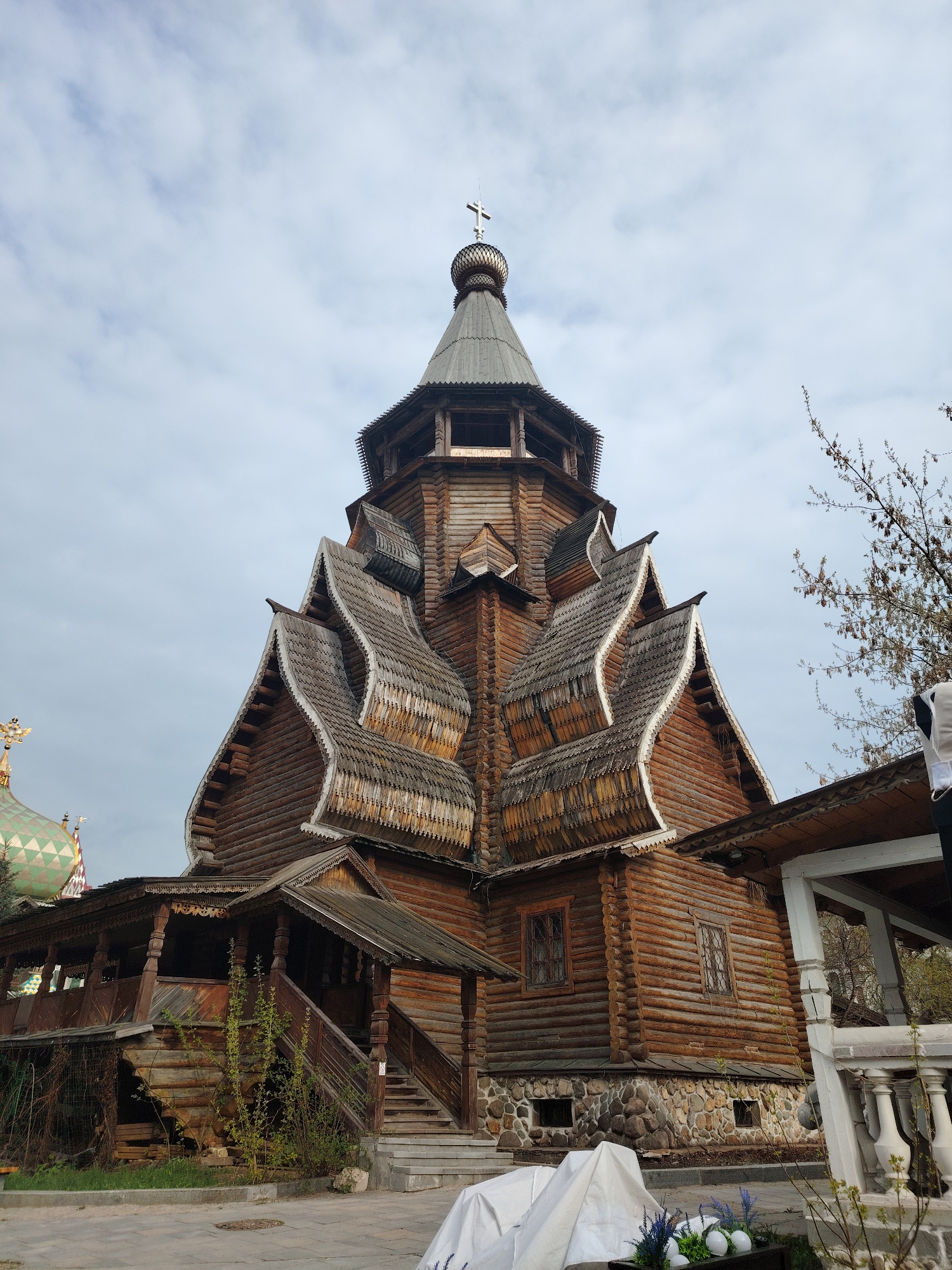 The image captures a striking, multi-tiered wooden building, likely an Orthodox church, characterized by its distinctive traditional Russian architecture. The structure is built from dark, horizontal logs and features a tall central tower with a pointed, shingled roof, topped by a small, gilded dome and an Orthodox cross. Below the tower, several intricately curved, shingled roofs, resembling "kokoshnik" gables, cascade downwards, showcasing detailed wooden craftsmanship. The base of the building rests on a robust stone foundation.
To the left, a portion of another building with a green, patterned onion dome and a cross is visible. On the right side of the foreground, part of a white structure with classical balusters can be seen, and a tree with bare branches extends into the frame.
The sky is overcast with light clouds, indicating daytime with diffused light. In the bottom foreground, a white tarp covers an object, and next to it, a dark planter holds white, egg-shaped decorations and small blue and purple flowers, possibly suggesting an Easter-related display or general decor. No people or specific activities are present, focusing solely on the architectural subject. The scene conveys a sense of historical and cultural significance, characteristic of sites in Moscow, Russia. No visible text is present.