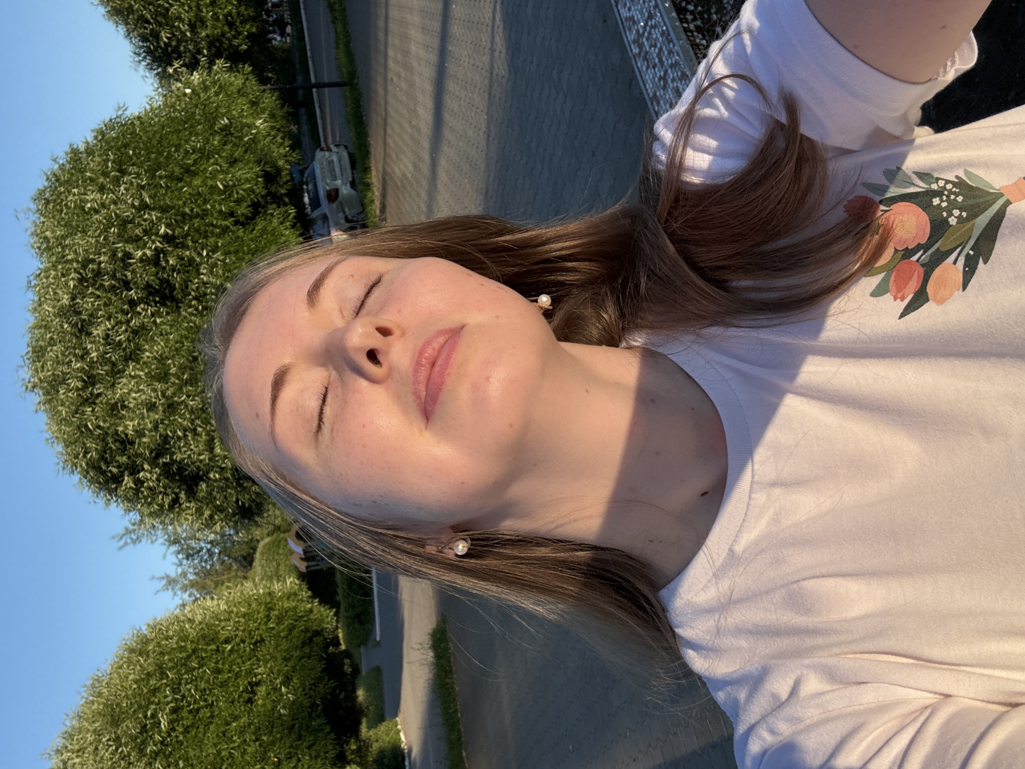 A young Caucasian woman with long, straight blonde hair is taking a selfie outdoors during the golden hour. She is wearing a white t-shirt with a floral print of tulips. Her eyes are closed, and her face is tilted upwards, bathed in warm sunlight. She is wearing pearl earrings. The background consists of lush green trees, a paved walkway, and a silver car parked in the distance. The setting appears to be a park or a residential area.