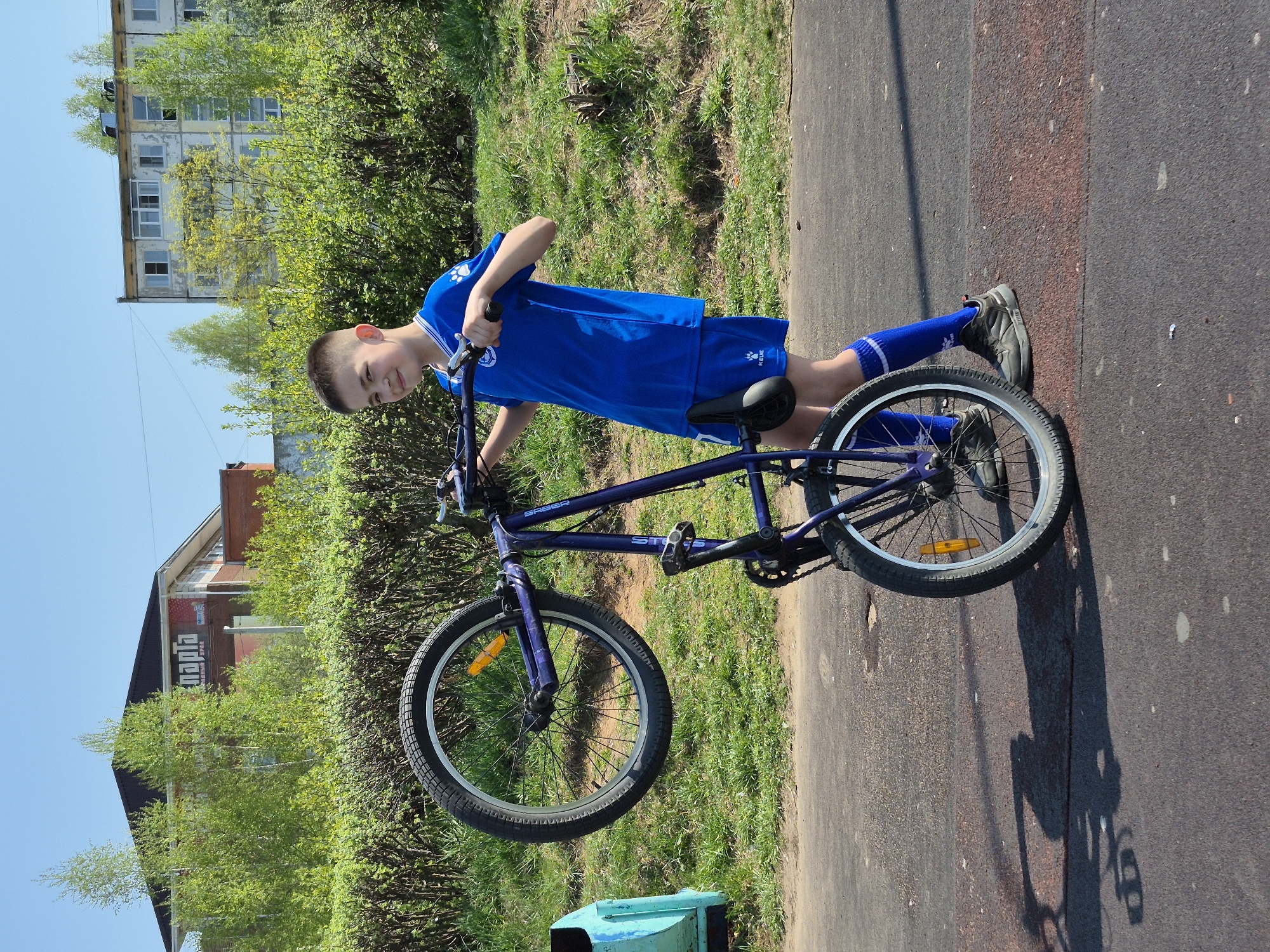 A young boy in a blue soccer uniform is standing on a paved surface next to a purple bicycle. He is holding the handlebars of the bike. The front wheel of the bike is slightly elevated off the ground. The background includes green bushes, grass, buildings, and a blue sky. The location is Chaikovsky, Russia.