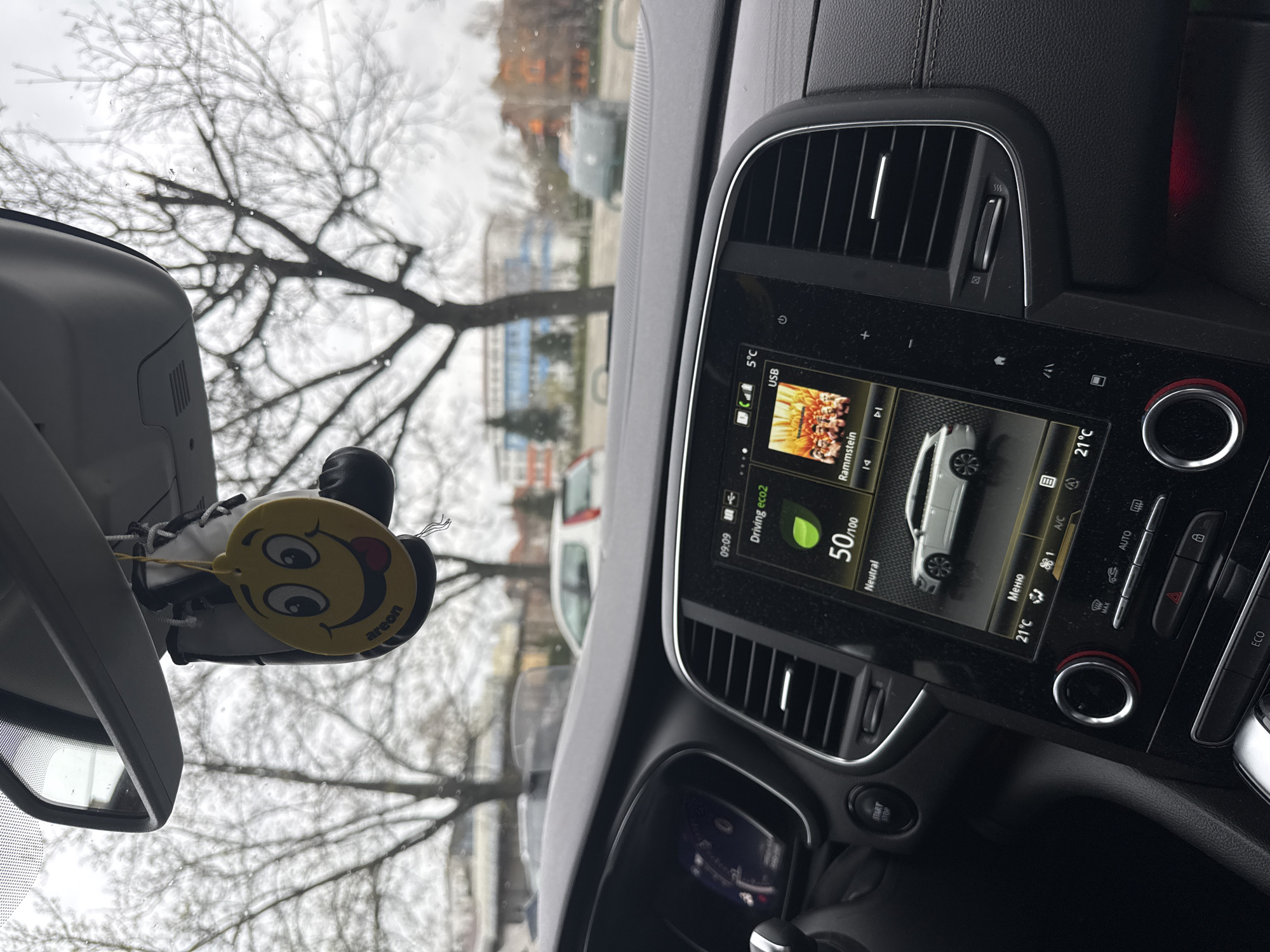 The image shows the interior of a car. The car is parked on a street in Panagyurishte, Bulgaria. A yellow air freshener with a smiley face and protruding tongue hangs from the rearview mirror. Through the windshield, bare trees and buildings are visible. The car's dashboard is in focus, displaying information such as the current temperature (5°C outside, 21°C inside), driving mode (eco2), and the current music being played (Rammstein). There is also a fuel efficiency score of 50/100. The car appears to be in neutral. Several control buttons and air vents are visible on the dashboard.
