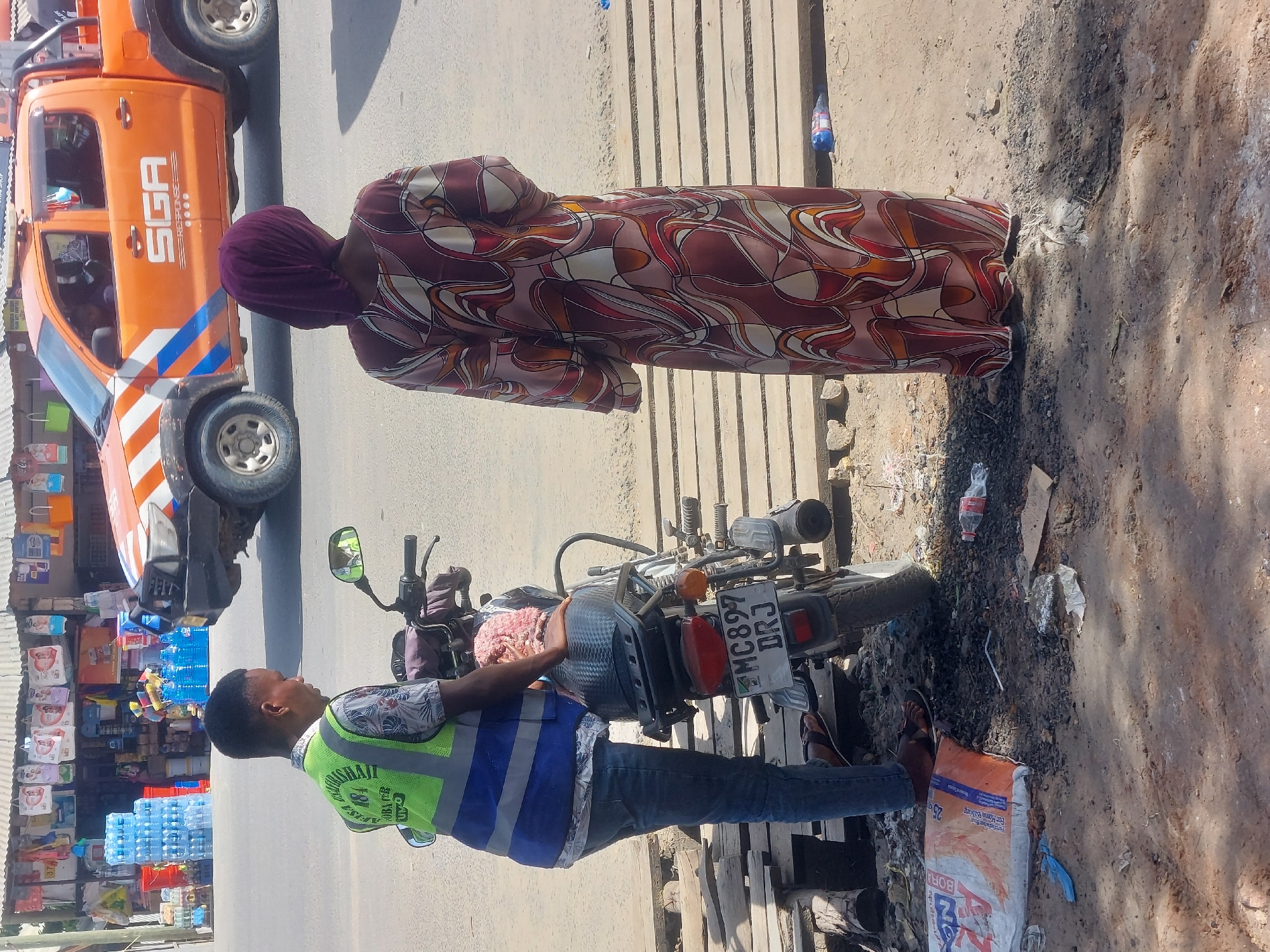 In Dar es Salaam, Tanzania, a roadside scene depicts two individuals and a parked motorcycle near a storefront. A woman wearing a maroon patterned dress and a purple head covering stands next to a man. The man is dressed in jeans, a patterned shirt, and a green and blue striped high-visibility vest. He is positioned next to a motorcycle with the license plate "MC897 DRJ." The motorcycle is parked on a rough, debris-strewn patch of ground next to some wooden planks. Behind the man and the motorcycle is a storefront with shelves stocked with various goods, including bottled water and other items. To the left, a portion of an orange vehicle is visible with the word "SGA" printed on it. The environment appears to be a busy street with buildings and other businesses in the background.