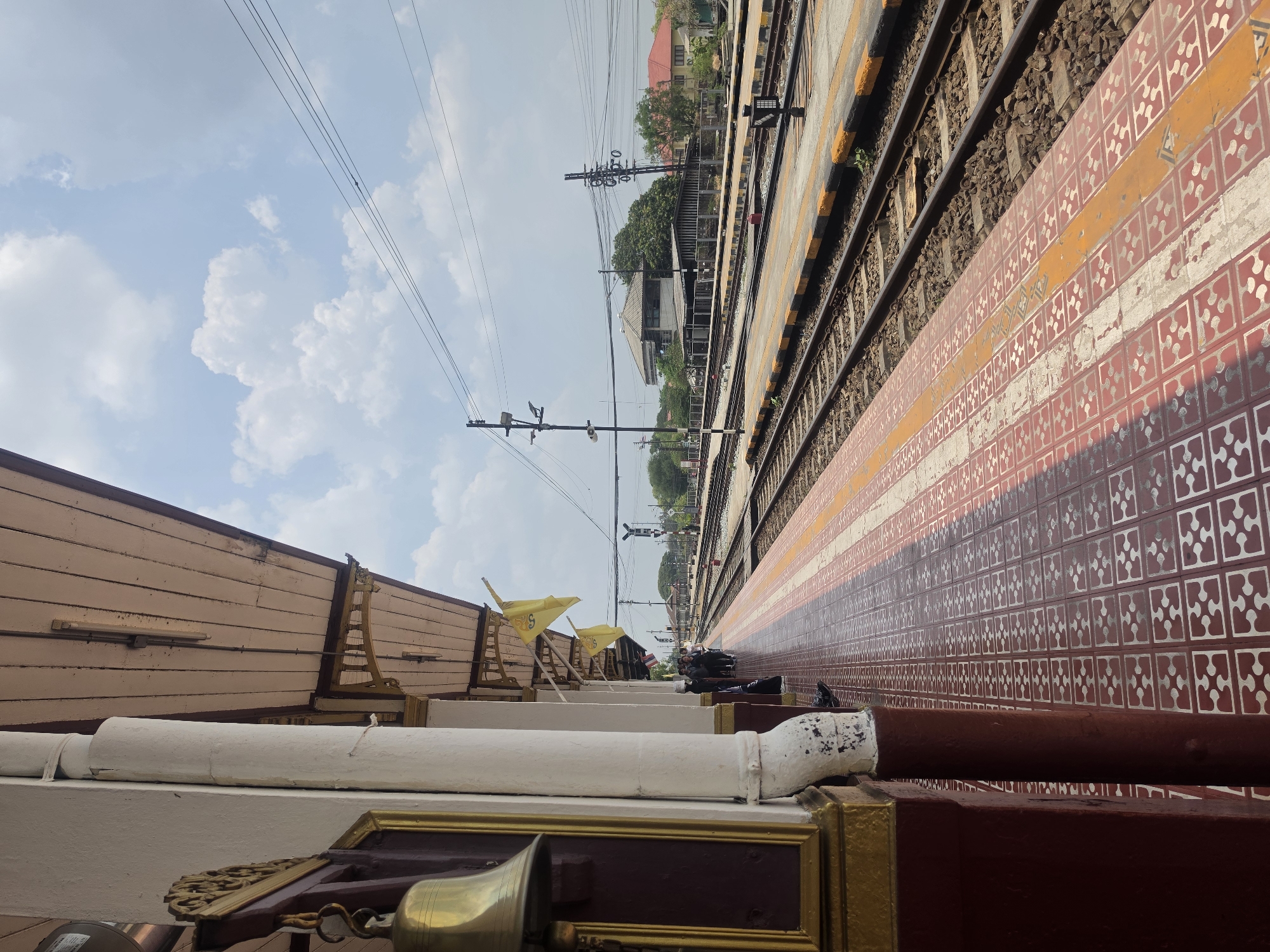 This image depicts a view from a train station platform in Phra Nakhon Si Ayutthaya City Municipality, Thailand. The platform is tiled with a patterned design in red, white, and orange hues. Railway tracks run parallel to the platform. Overhead, power lines stretch into the distance, juxtaposed against a partially cloudy sky. A wooden structure with white and gold trim is in the foreground, including a brass bell. Further down the platform, some figures are visible.