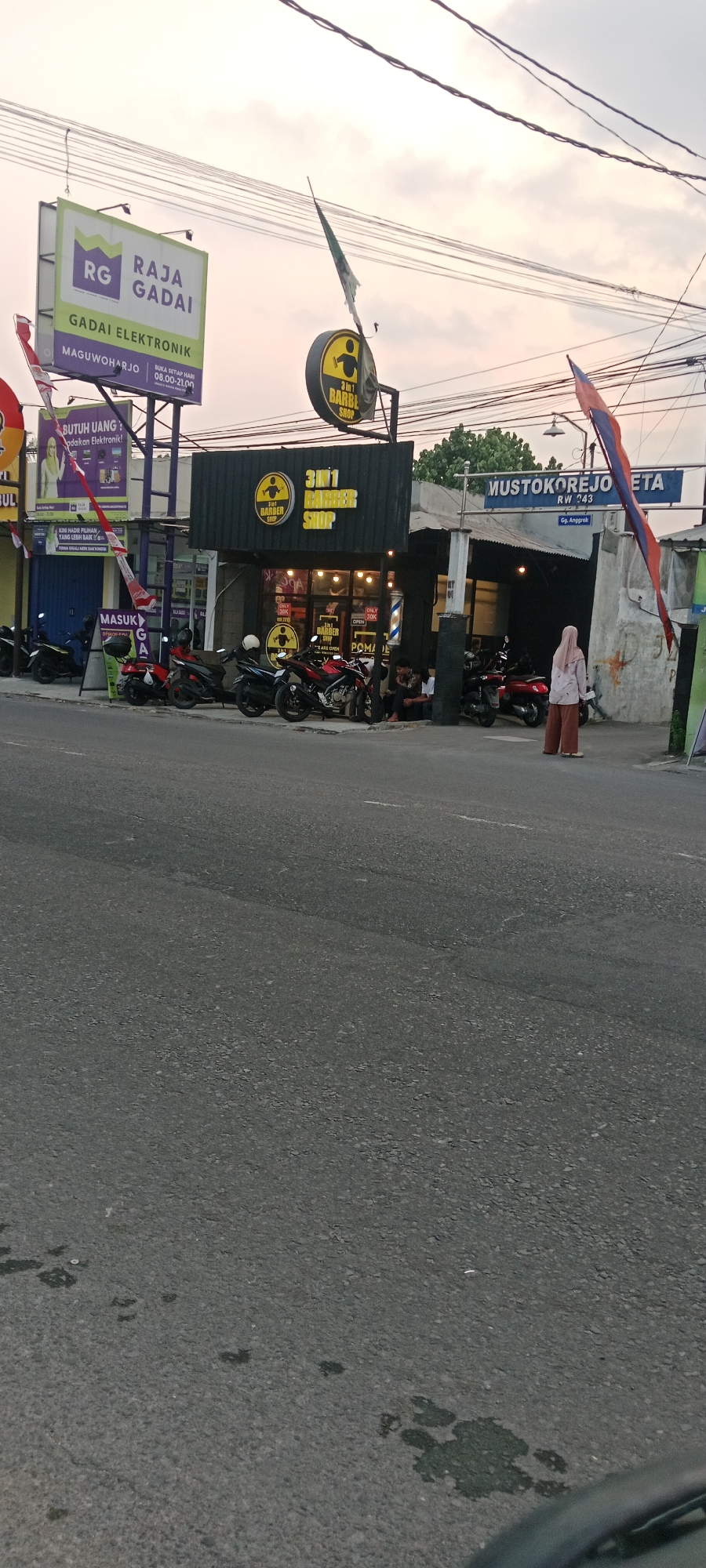 The image depicts a street scene in Maguwoharjo, Indonesia, at dusk. The foreground is dominated by a paved road with some oil stains. On the right side of the road, a woman wearing a hijab and loose-fitting pants stands facing away from the camera. Behind her, a street sign reads "MUSTOKOREJO RW 243" and "Gg. Anggrek". A white building with a grey pillar is visible.
The middle ground features several businesses. A prominent sign with the text "3 IN 1 BARBER SHOP" is displayed on a dark facade. The shop has large windows that reveal interior lighting and posters. Several motorcycles are parked in front of the barber shop. To the left of the barber shop, another establishment has a large green and white billboard for "RG RAJA GADAI GADAI ELEKTRONIK MAGUWOHARJO". A younger woman is visible in a poster on this billboard. Another sign in front of this establishment reads "MASUK GA". Additional motorcycles are parked further to the left.
The background shows power lines crisscrossing against a cloudy sky, suggesting the approach of evening. Trees are visible in the distance. Flags, including a tattered Indonesian flag, are attached to poles. The overall environment appears to be a commercial area.