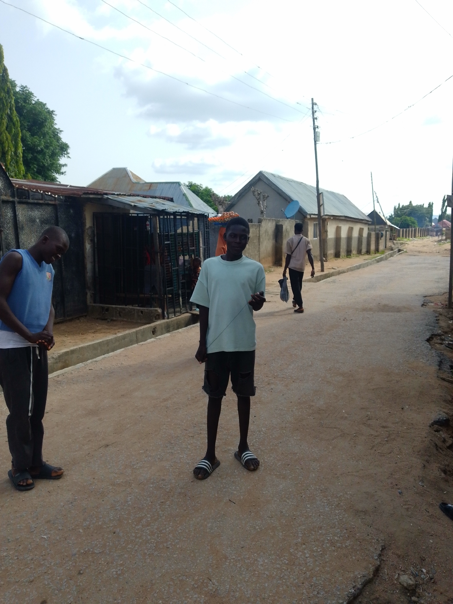 The image depicts an outdoor scene on a paved street in Jalingo, Nigeria. Two young men are the primary subjects, standing on the street. The man on the left wears a blue sleeveless t-shirt over a white t-shirt and dark pants. He is looking down and to his left, with a slight smile. The man on the right is wearing a light blue t-shirt and dark shorts. He is facing the camera, holding a thin string in his right hand. In the background, another man walks away from the camera, carrying a bag.
The setting includes buildings with corrugated metal roofs lining the street, suggesting a residential or commercial area. There are also power lines and a utility pole visible. The ground is a mix of dirt and pavement, with some areas appearing worn or damaged. A tall, dense green tree is visible on the far left. The sky is cloudy, indicating possibly overcast weather. The overall mood appears casual and unposed, capturing a moment in everyday life in the location. No visible text is present in the image.