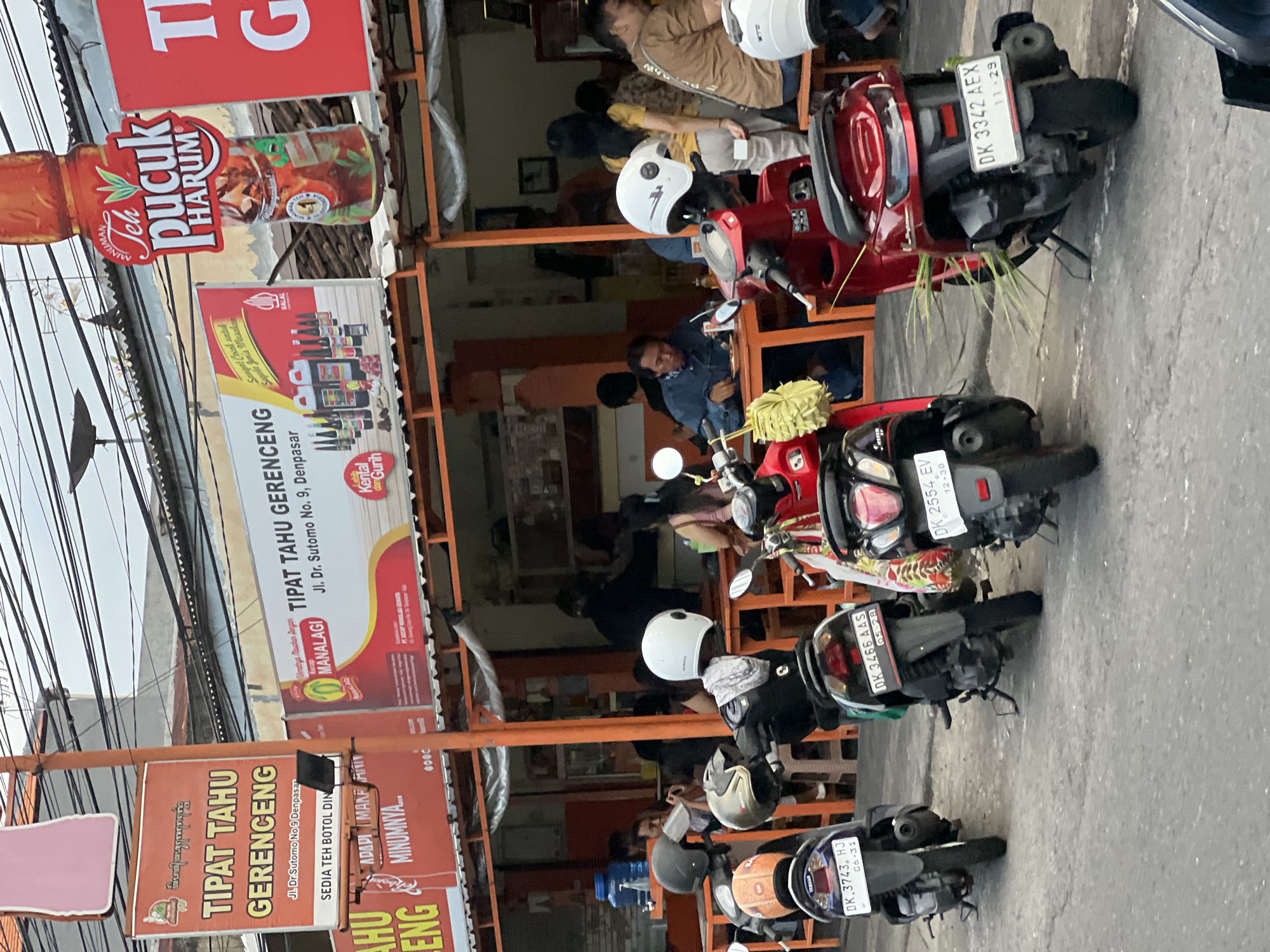 This is a street-level photograph taken in Denpasar, Indonesia, likely depicting an outdoor dining establishment with motorcycles parked in front. The scene features several scooters and motorcycles parked along a paved street, some with helmets placed on them. In the background, there is an open-air restaurant with orange support poles and awnings. Several people are seated at tables inside the restaurant, some appearing to be eating or engaged in conversation. Signs for "TIPAT TAHU GERENG" and "Teh Pucuk Harum" are prominently displayed, indicating local food and beverage businesses. The overall environment suggests a moderately busy urban setting.