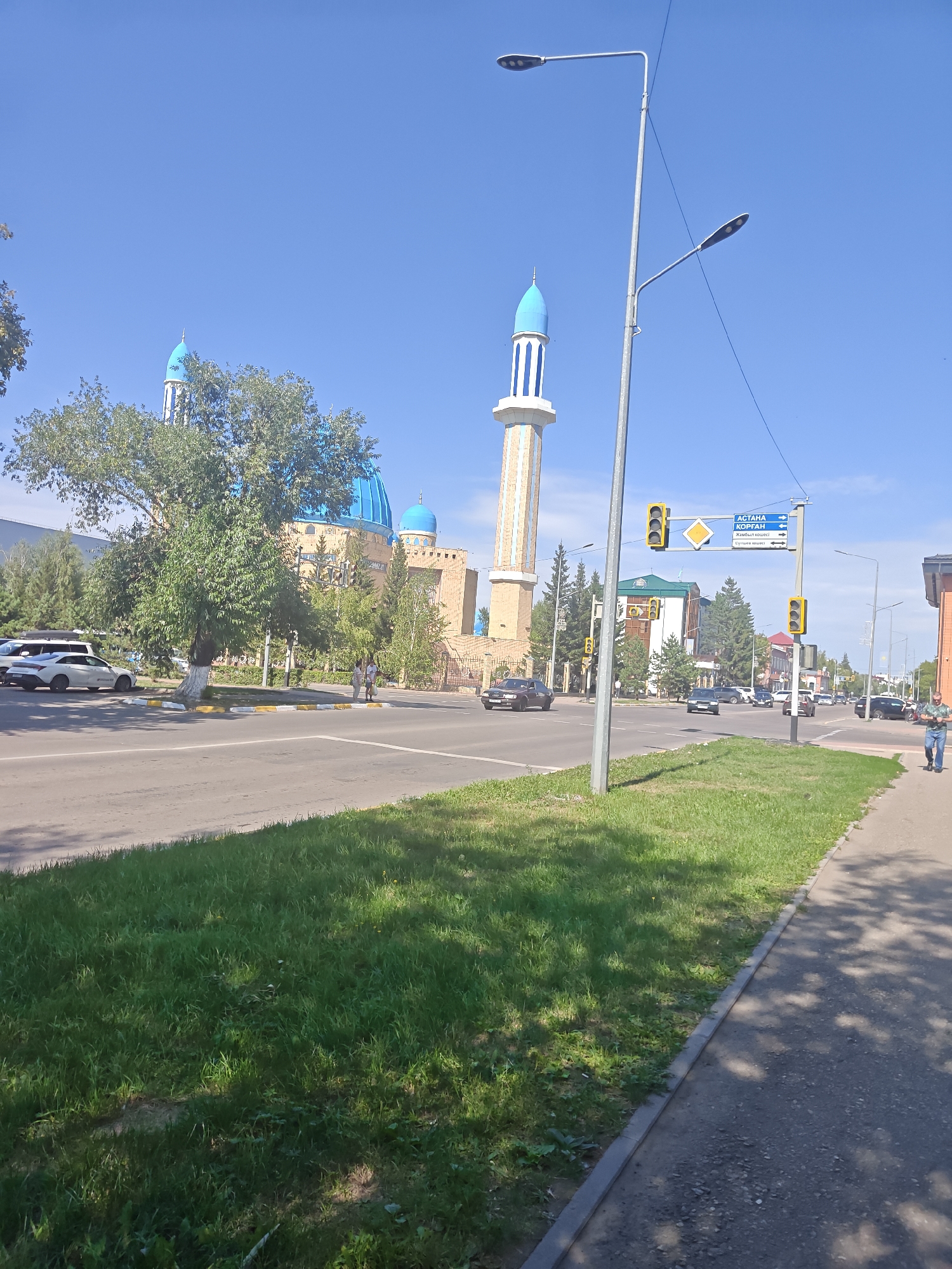 The image shows a street scene in the city of Petropavl, Kazakhstan, on a clear, sunny day. A prominent mosque with blue domes and a tall minaret dominates the background. The mosque is an ochre color with decorative elements on the minaret. In the foreground, a paved road runs horizontally across the image, with a grassy median strip and a sidewalk on the right. Several vehicles, including cars and what appears to be an SUV, are visible on the road. A traffic light with a diamond-shaped sign is positioned near the intersection. Two streetlights are also visible. A few pedestrians are present on the sidewalk and crossing the street. Trees and other vegetation line the roadside. A signpost with text in Cyrillic script indicates directions to "АСТАНА", "КОРГАН", and "Жамбыл көшесі". The overall environment suggests an urban setting with elements of religious architecture and daily life.