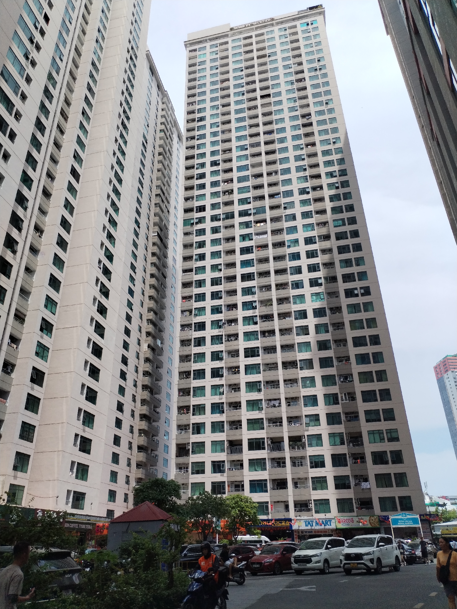 In a low-angle, wide shot, three tall, light-colored apartment buildings dominate the frame, with the building on the right angled and leaning towards the viewer. The buildings are densely packed with numerous windows, some open, and balconies displaying laundry. Below the apartment buildings, a street scene unfolds with several vehicles, including cars and motorcycles, parked and moving. Pedestrians are visible on the sidewalk and street. Green trees and foliage partially obscure the lower portions of the buildings and street. Signs are visible on the buildings and street, indicating commercial establishments. The sky is overcast. The scene appears to be in a densely populated urban environment.