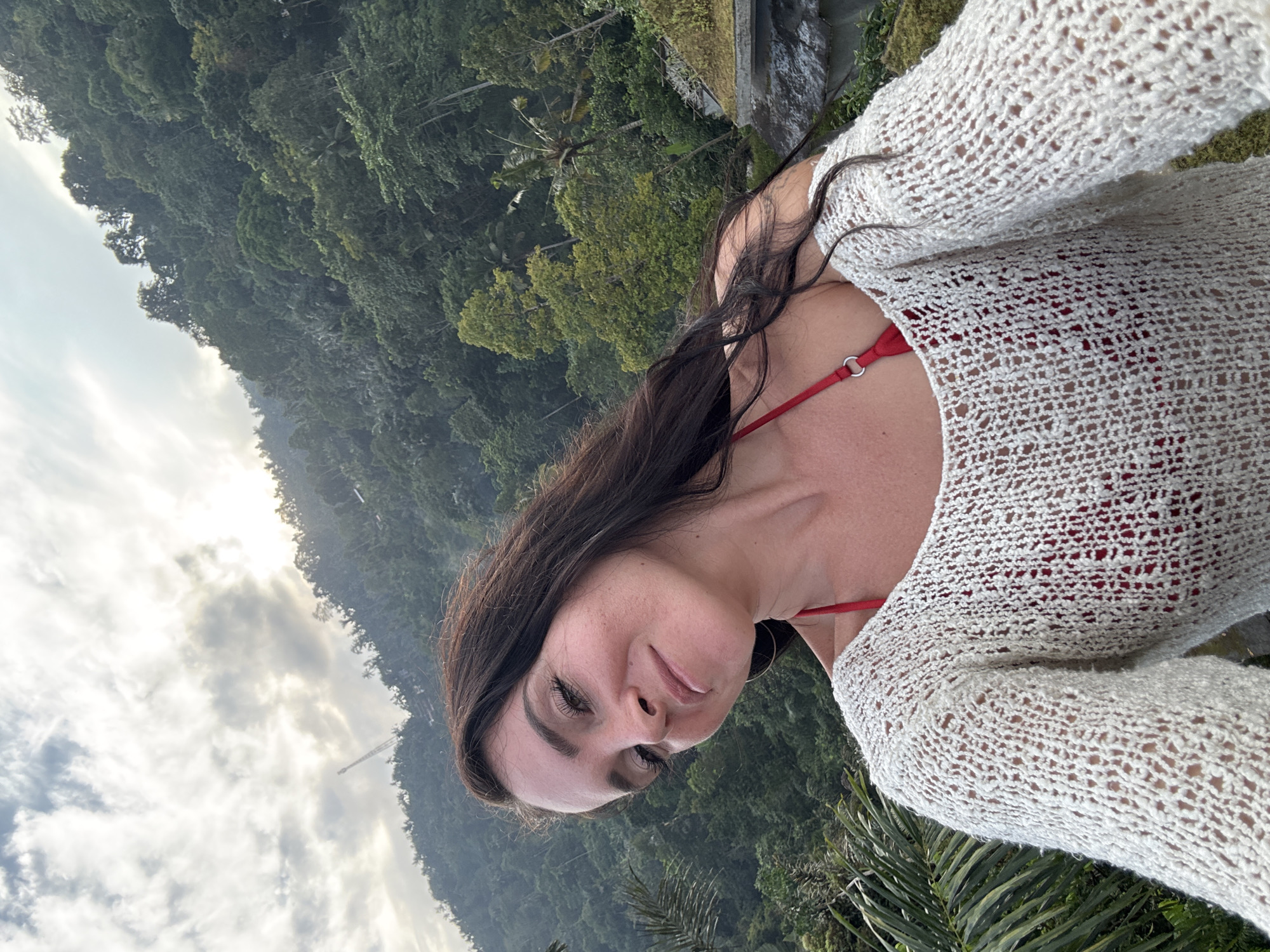 A woman with dark brown hair takes a selfie in front of a lush, green mountainous landscape. She is wearing a red bikini top and a cream-colored, open-knit sweater. The background features dense jungle foliage, with a cloudy sky above. The scene appears to be outdoors in a tropical environment, likely near Bebandem, Indonesia, given the terrain and vegetation.
