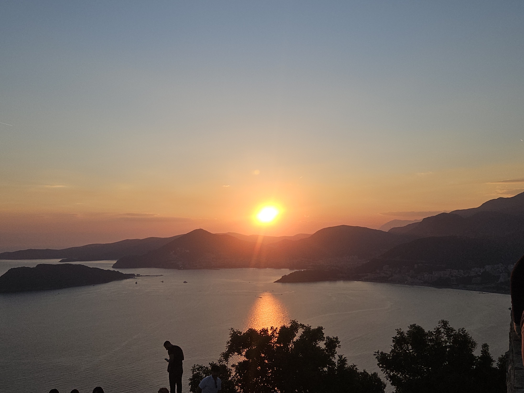 The image captures a sunset over the Adriatic Sea, with the island of Sveti Stefan visible in the distance. The sky is a gradient of soft blues and oranges, with the sun a bright orb near the horizon, casting a shimmering reflection on the water. Silhouetted against the vibrant sky, several people are standing on a hill, overlooking the scene. One person in the foreground is looking down at their phone, while others are gathered in a group. Lush green trees frame the bottom of the image, adding a natural element to the picturesque view.