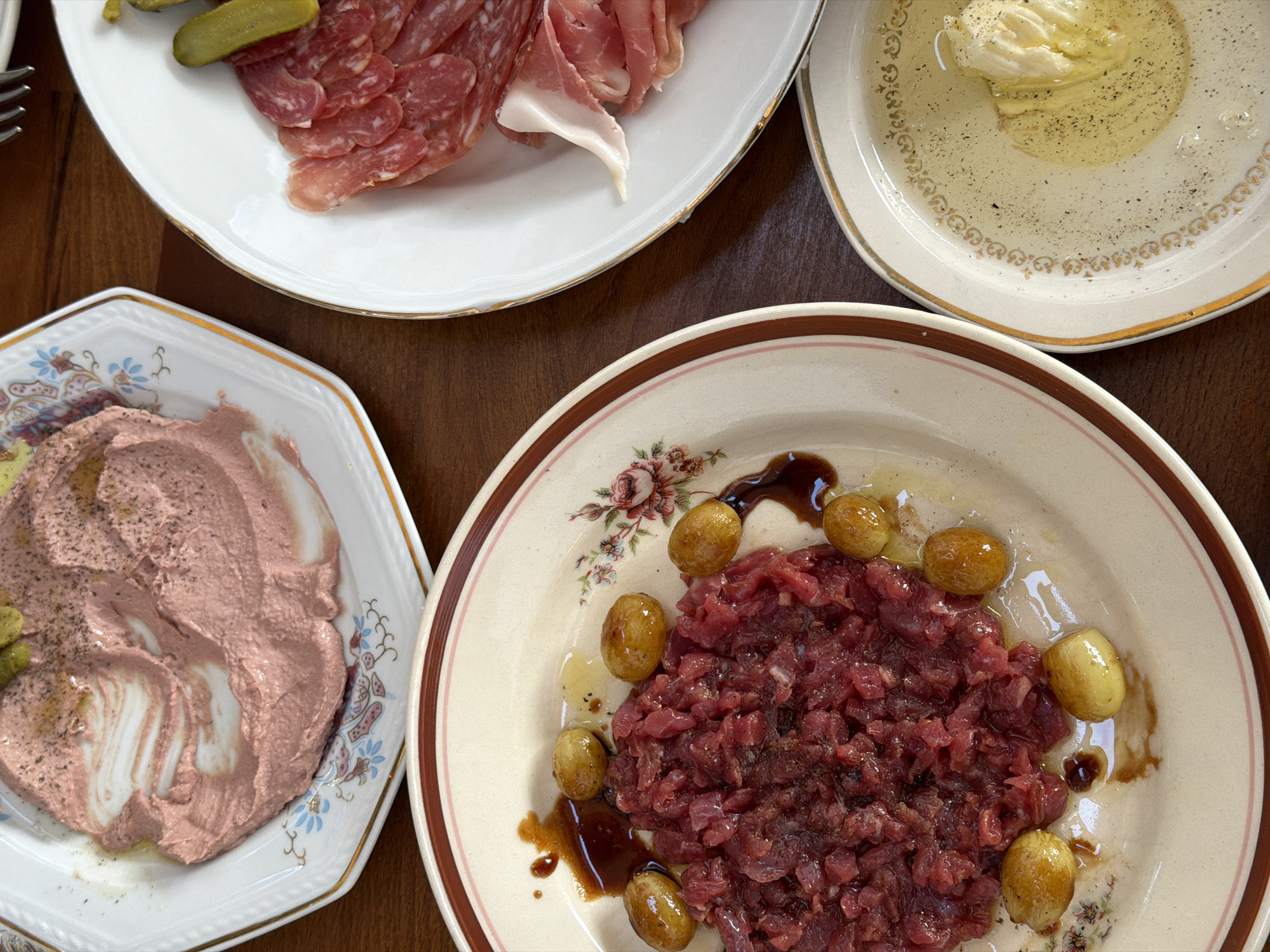 The image shows a close-up overhead view of a wooden table set with several plates of food, likely a meal in Belgrade, Serbia. The lighting is dim and warm, suggesting an indoor setting such as a restaurant or home.
In the foreground and to the left, a round, cream-colored plate with a floral design features a mound of finely diced raw red meat, likely beef tartare, garnished with small, golden-brown roasted potatoes and drizzled with a dark sauce.
To the right of the tartare, another plate, also cream-colored with gold trim and decorative patterns, holds a selection of cured meats, including thinly sliced salami and prosciutto, along with a gherkin.
Further to the left, another cream-colored plate with a blue and floral pattern contains a light pinkish-brown spread, possibly a pâté or dip, with visible swirls and a dusting of pepper. Another gherkin is visible on this plate.
At the bottom right, a small, decorative plate holds a dollop of what appears to be burrata or a soft cheese, bathed in a clear, yellowish liquid, likely olive oil, and seasoned with pepper.
The overall impression is of a shared meal with several appetizers or small plates, presented on vintage-style tableware. The focus is on the food, with the background showing the dark wood grain of the table. No people are visible in the image.