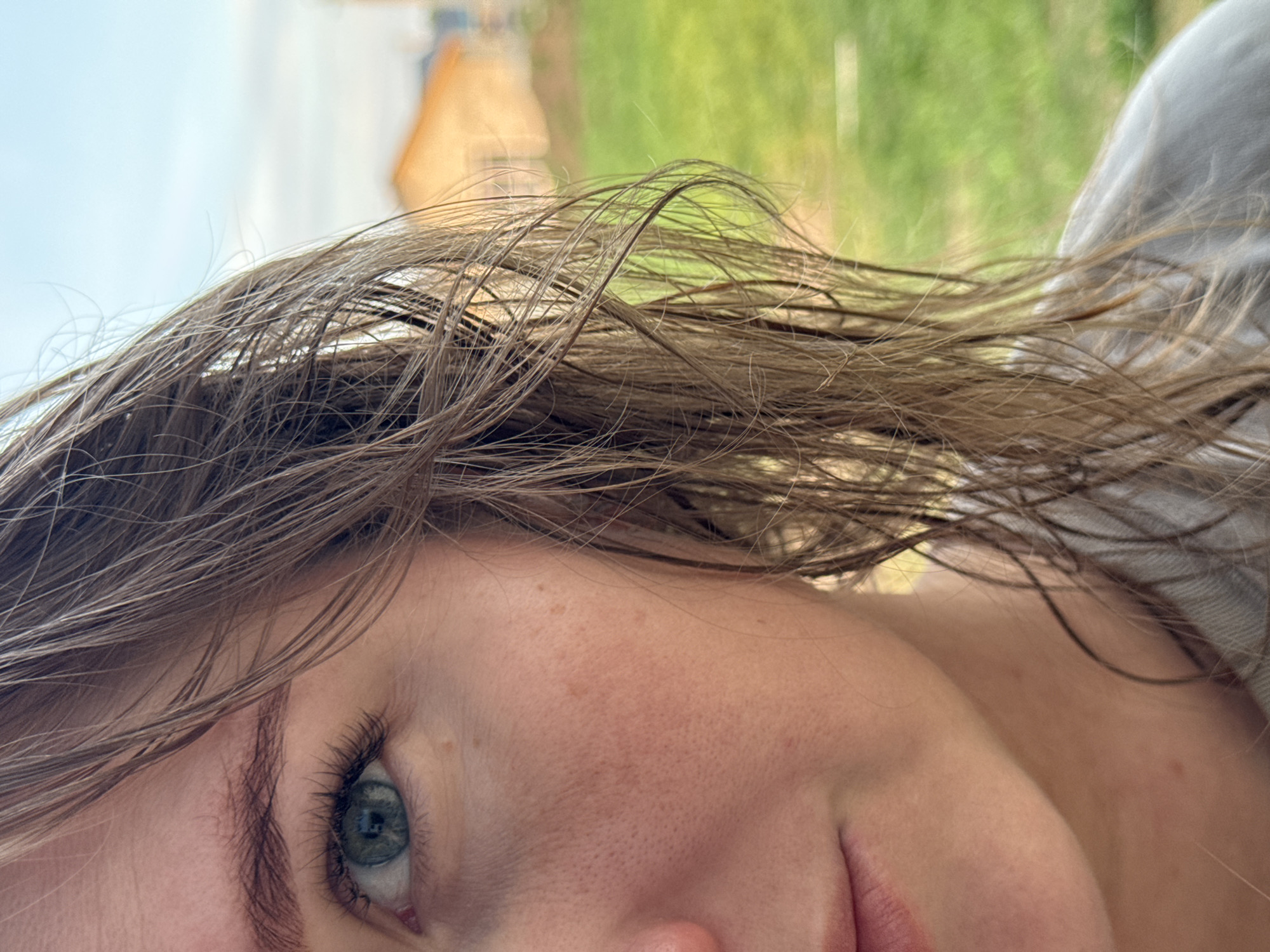 Close-up angled view of a Caucasian woman's face, appearing to be outdoors. Her hair is wet and brown, partially obscuring her face. She has fair skin, visible freckles, and green eyes. A portion of her grey clothing is visible on the right edge of the image. The background appears to be a mix of blurred greenery and what appears to be part of a building. Given the location is Ферма, Russia, the style of building may indicate a rural or suburban environment. There are no discernable actions or interactions occurring in the frame.
