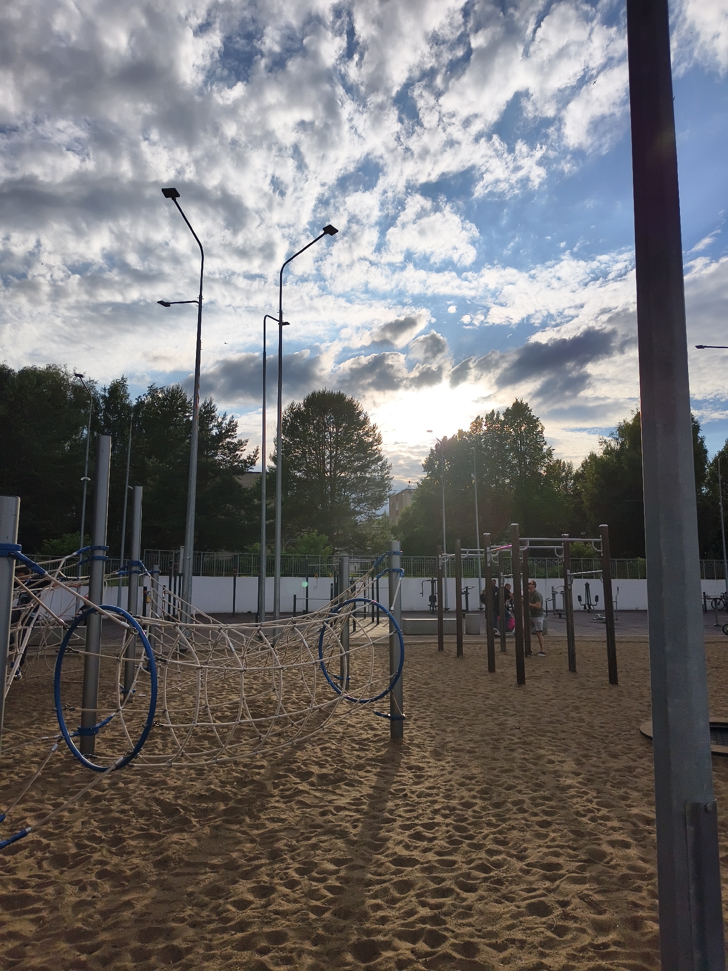 This is a wide-angle outdoor shot of a public park with a playground and outdoor exercise equipment. The ground is covered in sand, and in the foreground, a rope tunnel structure with blue rings is visible. In the middle ground, there are several pieces of exercise equipment, including pull-up bars and weight machines. A few people are scattered around the exercise area, engaged in or observing activities.
Behind the exercise equipment, a low fence separates the park from a tree-lined area with buildings in the distance. The sky is dynamic with a mix of dramatic clouds and patches of blue sky, suggesting it might be late afternoon or early evening. Sunlight filters through the clouds, casting a warm glow on the scene. Tall lamp posts are positioned throughout the park, ready for illumination as dusk approaches. The overall atmosphere is calm and relaxed, indicating a typical day at a community park. The image appears to be taken in Chaikovsky, Russia. There is no visible text in the image.