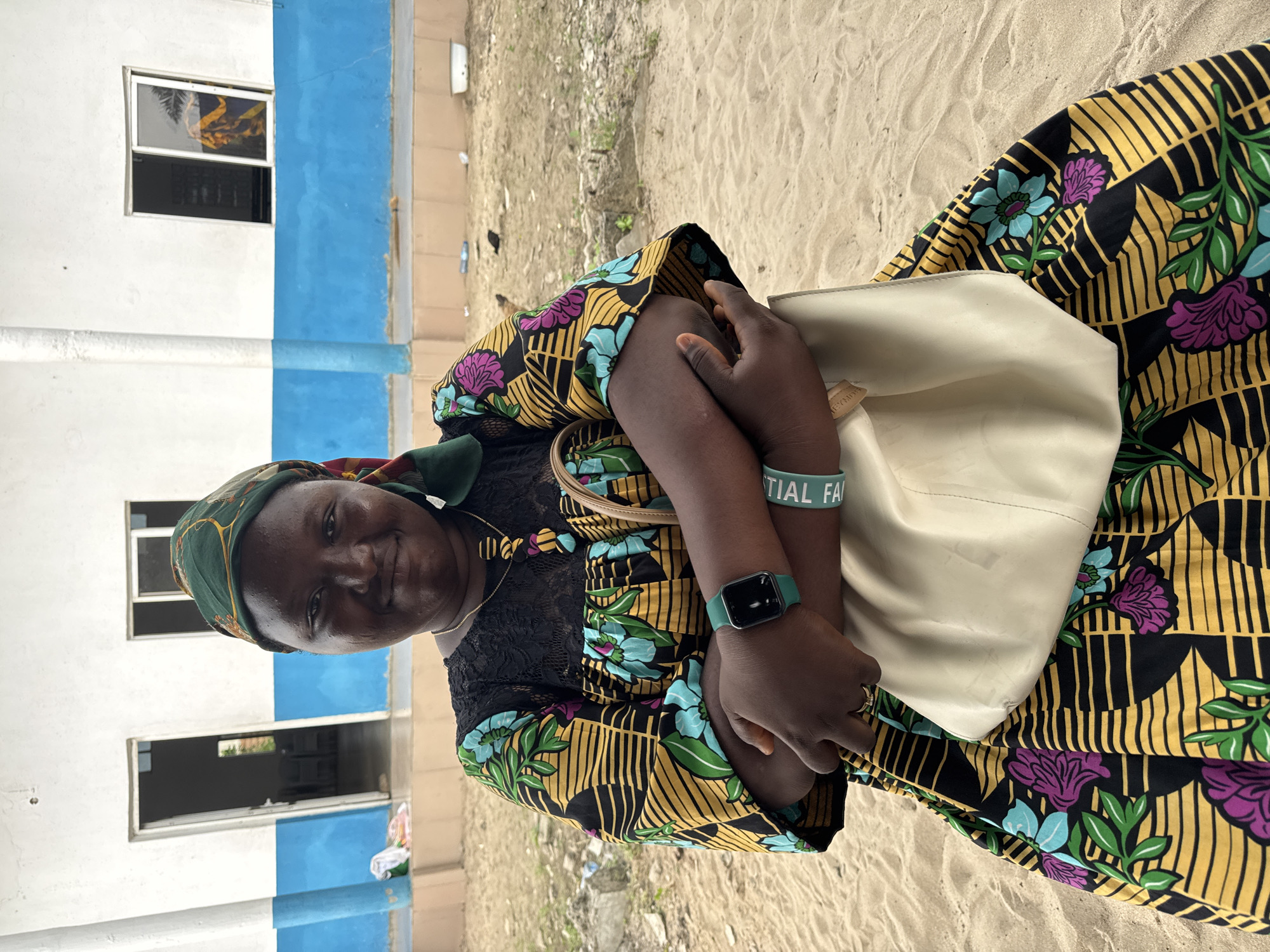 A woman stands outside a building in Badagry, Nigeria. The building is light colored with a horizontal band of blue. The woman wears a green headscarf, a patterned yellow, black, and floral dress, and a green wristwatch. She is holding a beige handbag. The ground is sand.
