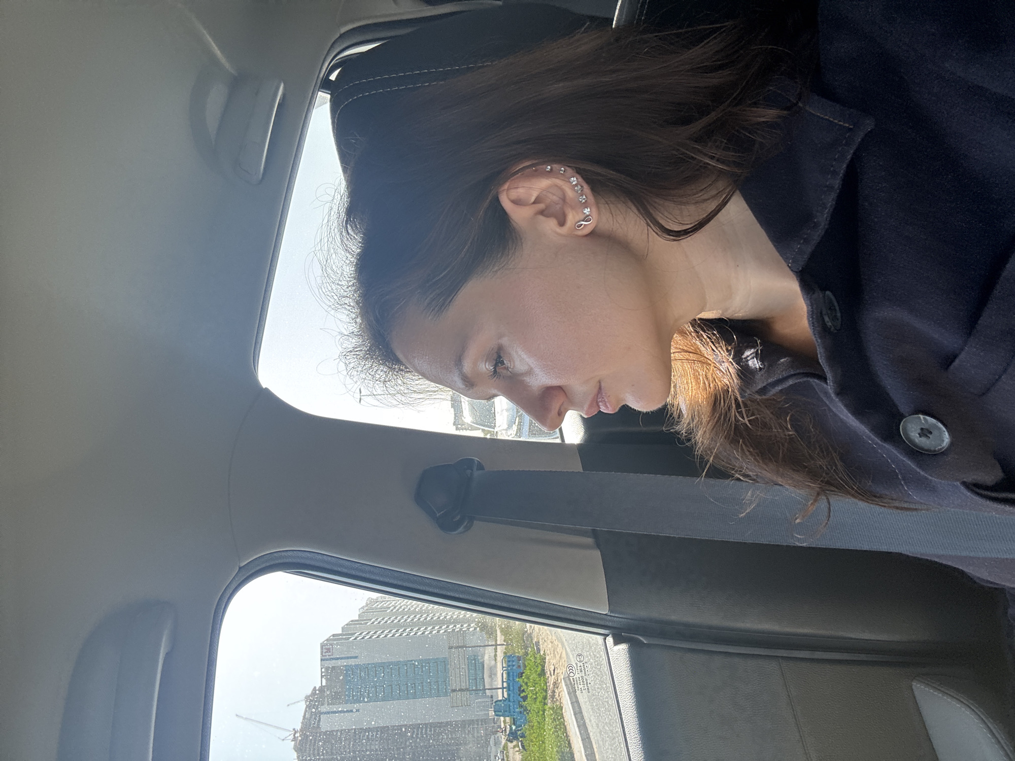A medium indoor shot, taken from a car interior, shows a woman sitting in the passenger seat, wearing a black jacket. Her face is turned downward, and her right ear, adorned with multiple earrings, is visible. Outside the car window, a cityscape is visible, featuring tall buildings, likely under construction, and green foliage. The location is Dubai, United Arab Emirates. The scene suggests the woman is either pensive or looking at something in her lap. A seatbelt is visible.