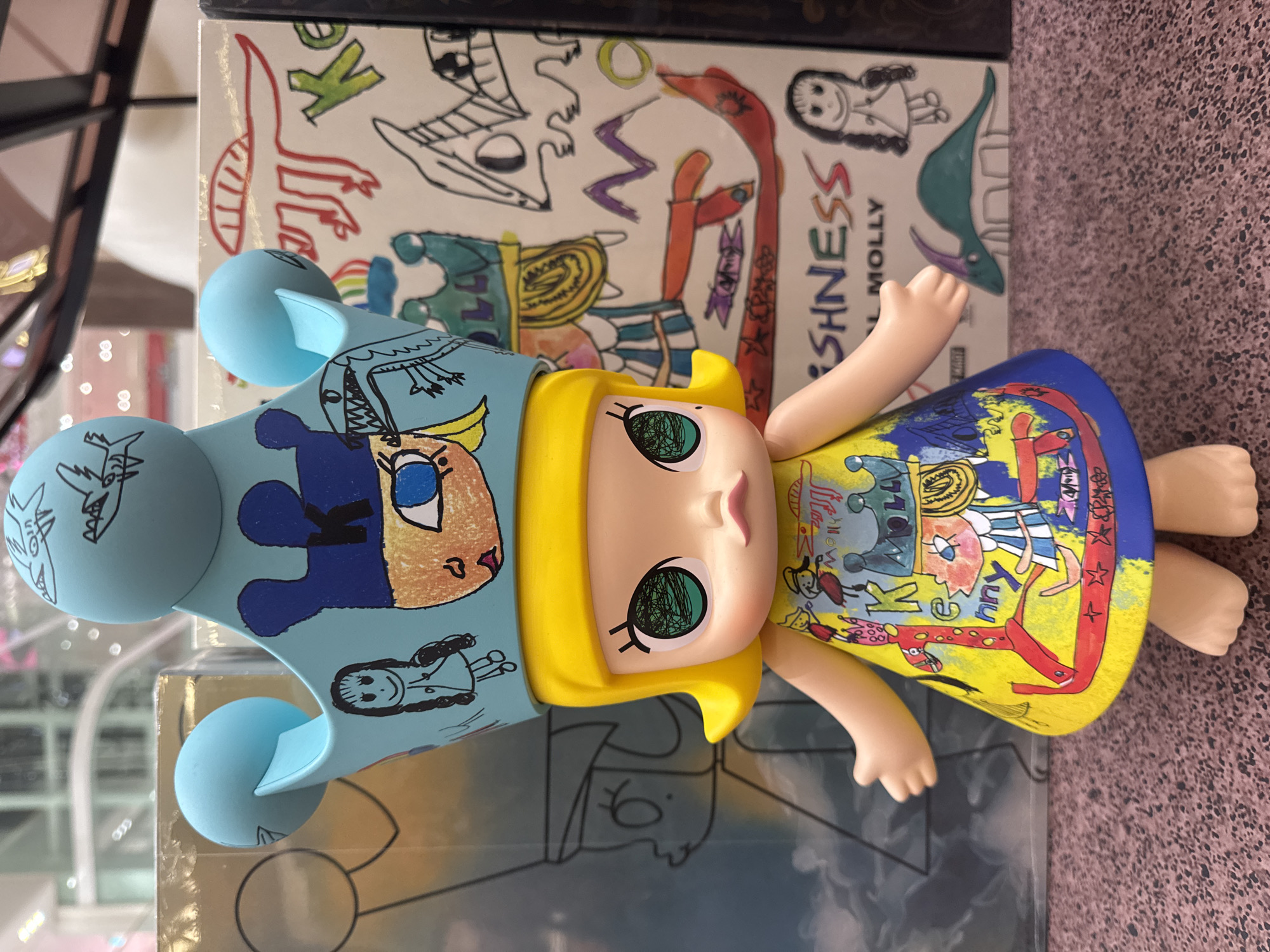 This media file captures a close-up, high-angle shot of a collectible doll, likely a "Molly" figurine from the brand "Freshness," given the visible text. The doll has bright yellow hair, large green eyes with scribbled pupils, and pale skin. It is wearing a blue headdress adorned with cartoonish drawings, including a crowned face and various characters. The doll's yellow dress is decorated with vibrant, childlike drawings in red, blue, and black, featuring stars and figures. The doll is positioned horizontally and appears to be resting on top of a box or book with a white background featuring more hand-drawn sketches in red, black, and purple. The overall aesthetic is colorful and playful. The background is blurred, but hints of a retail or display environment can be seen, with glass and possibly other items on shelves. The lighting suggests an indoor setting, likely during the day. The context of "Hà Nội, Vietnam" implies this item is located in that city.