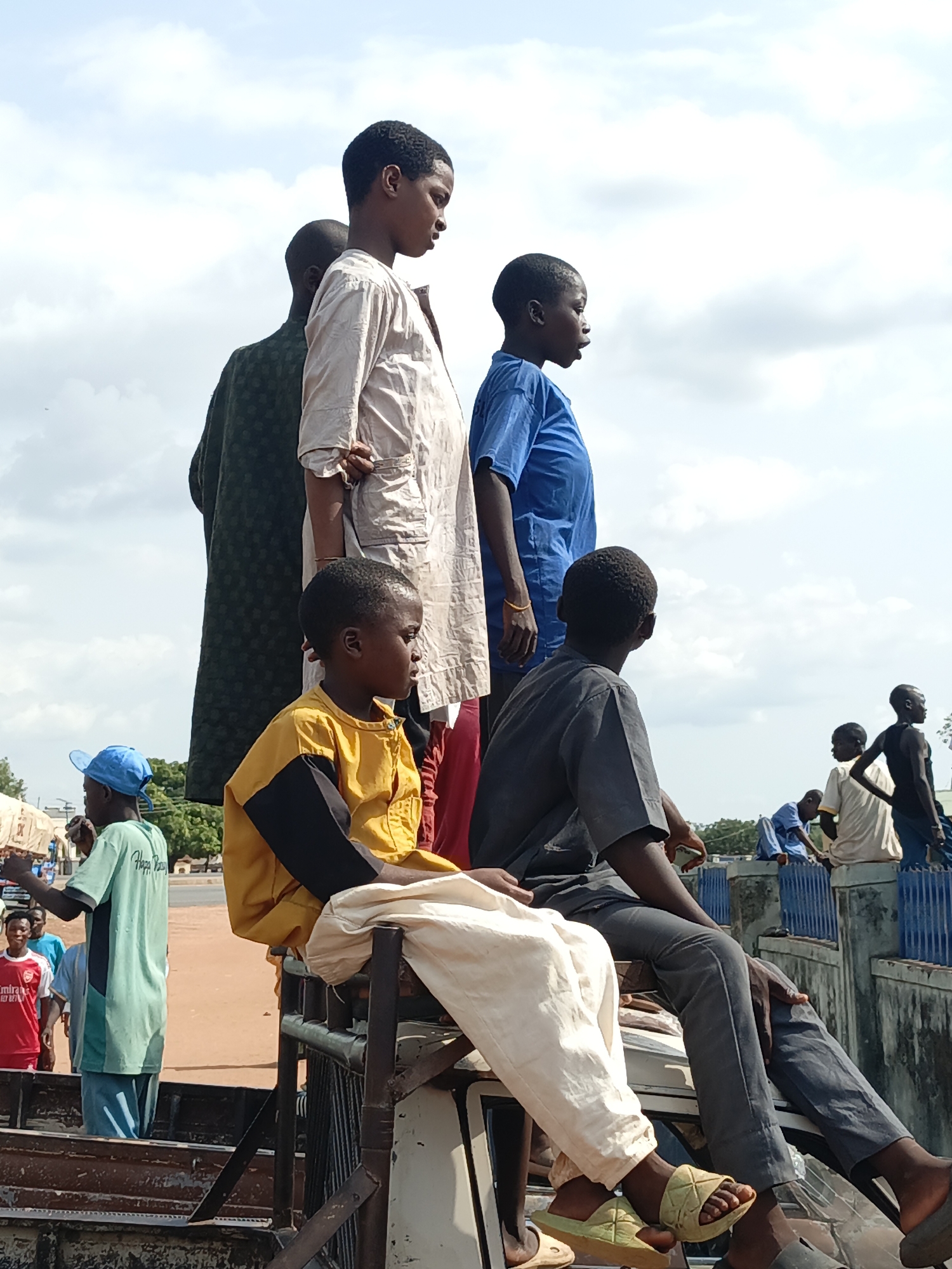 The image depicts a group of young people, primarily boys, riding in the back of a truck or vehicle in an urban setting. The sky is partly cloudy.
A young boy in a yellow and black jacket sits prominently on a metal structure. Beside him, a boy in grey pants and a grey shirt is sitting on a raised part of the truck. Other individuals are standing in the back of the truck, including a boy wearing a light-colored tunic and another in a blue shirt. A person in a dark patterned robe is also present.
In the background, other people and vehicles are visible, indicating an active street scene. A blue fence is seen. A boy in a red shirt and a boy in a green shirt and blue cap are visible as well. The scene appears to be taken during daylight hours in Lafia, Nigeria, based on available data and image characteristics.