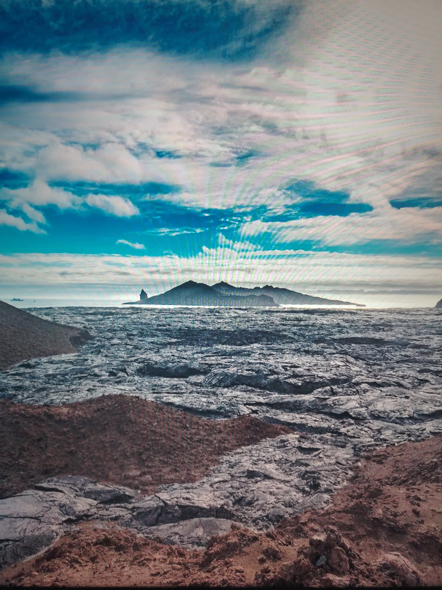 The image depicts a landscape with a rocky foreground composed of grey and brown textures. In the distance, across what appears to be a body of water, lies a mountainous landmass with a few distinct peaks. The sky is partially obscured by clouds, with patches of blue visible. The location appears to be a coastal or island environment, with no clear indication of human presence or specific urban elements. There is no mention of Unknown city, Unknown in the image.
