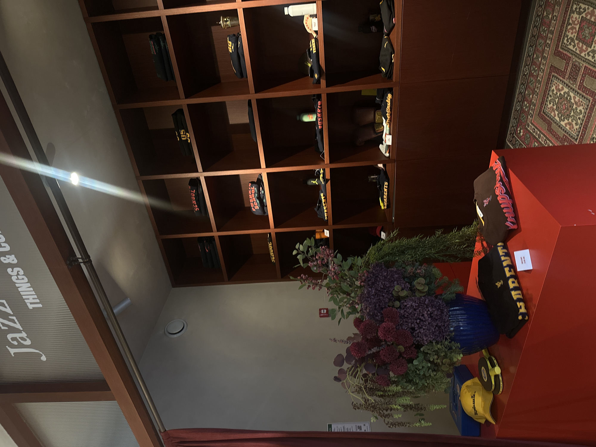 The image displays the interior of the "Jazzve Things & Coffee" cafe in Yerevan, Armenia. A large wooden shelving unit is seen filled with items for sale, including folded black t-shirts and black hoodies. In the forefront, a blue vase contains a large bouquet of flowers set on top of a red display counter. A black hat and a yellow hat sit next to the vase. The floor is covered in a patterned rug.