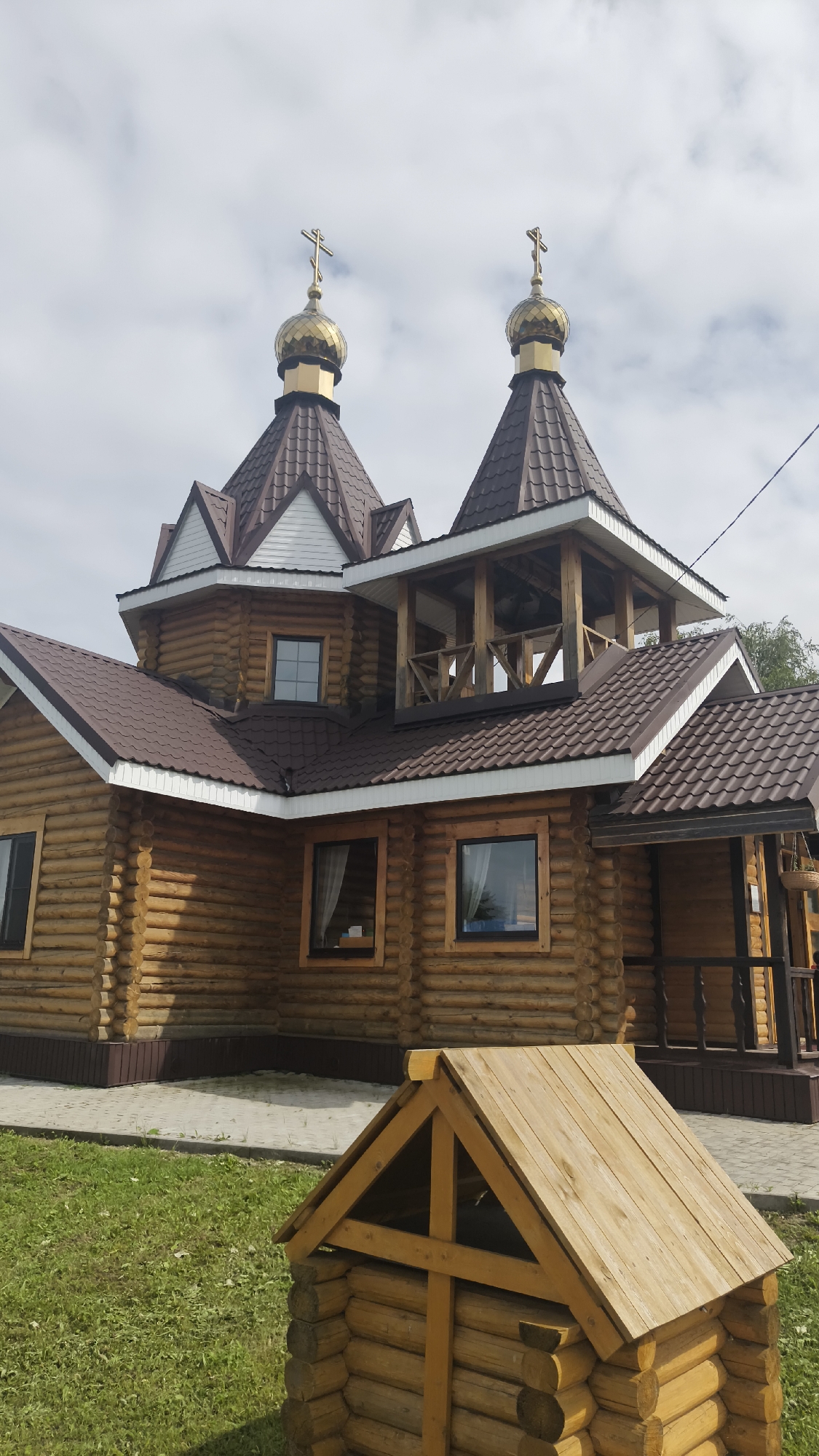The image shows a wooden building with multiple gabled roofs and two golden onion domes topped with crosses, indicative of an Orthodox church. The structure is constructed from horizontal logs. There are several windows visible, some with curtains. In the foreground, a small, wooden, gabled structure made of logs is situated on a patch of green grass. The background consists of a cloudy sky and some trees. The scene is located in the village of Stanovaya, Russia.