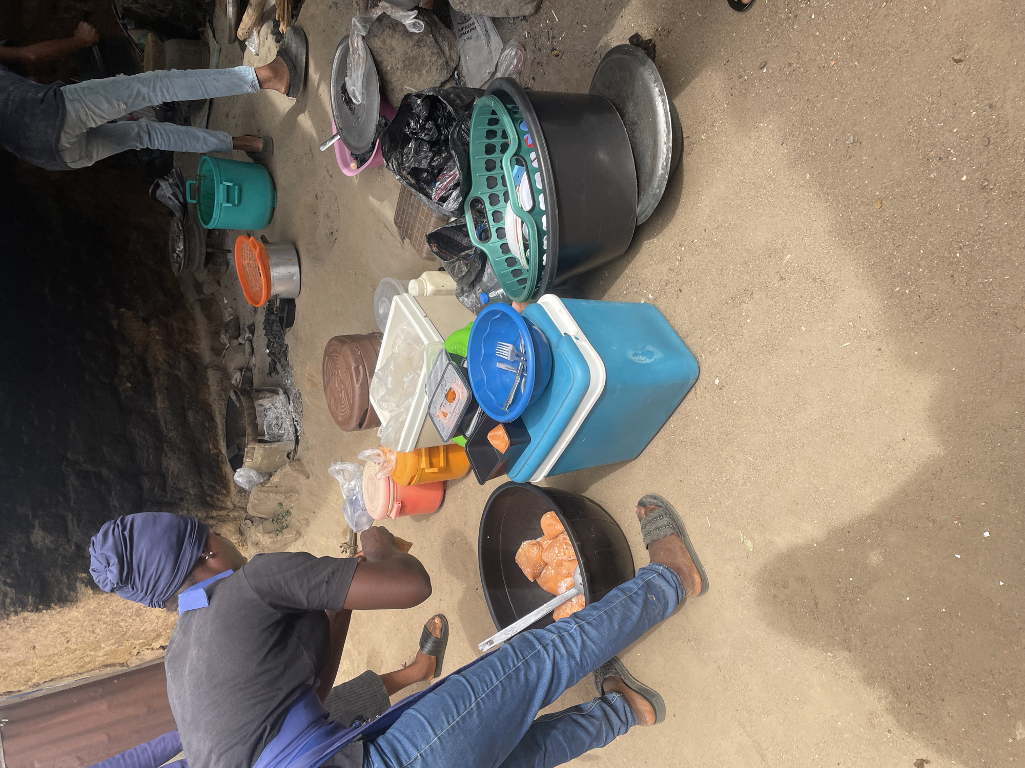 The image captures an outdoor scene of food preparation on sandy ground, likely in Jos, Nigeria. In the foreground, a person wearing a dark shirt, blue headwrap, blue apron, blue jeans, and sandals is seated or crouching. They are actively engaged with a large black plastic tub containing numerous orange-colored food items, holding a pair of tongs.
To the left, another person's lower legs and feet are visible, standing near a traditional cooking area. This area features an earthen structure with a dark opening, suggesting a fire pit or cooking stove, surrounded by ash and stones.
Various plastic containers and kitchenware are arranged around the space. These include a large blue cooler, a blue bowl filled with metal utensils (forks, spoons), a green plastic basket, a green plastic bucket, an orange plastic colander, and several smaller containers and plastic bags. A large metal pan or lid is also visible in the background. On a white bag in the mid-ground, the word "GURUM" is partially visible.
The scene depicts a casual, active food handling or cooking environment during the daytime, indicated by shadows on the dry, sandy ground. The overall impression is one of communal meal preparation in a local setting.