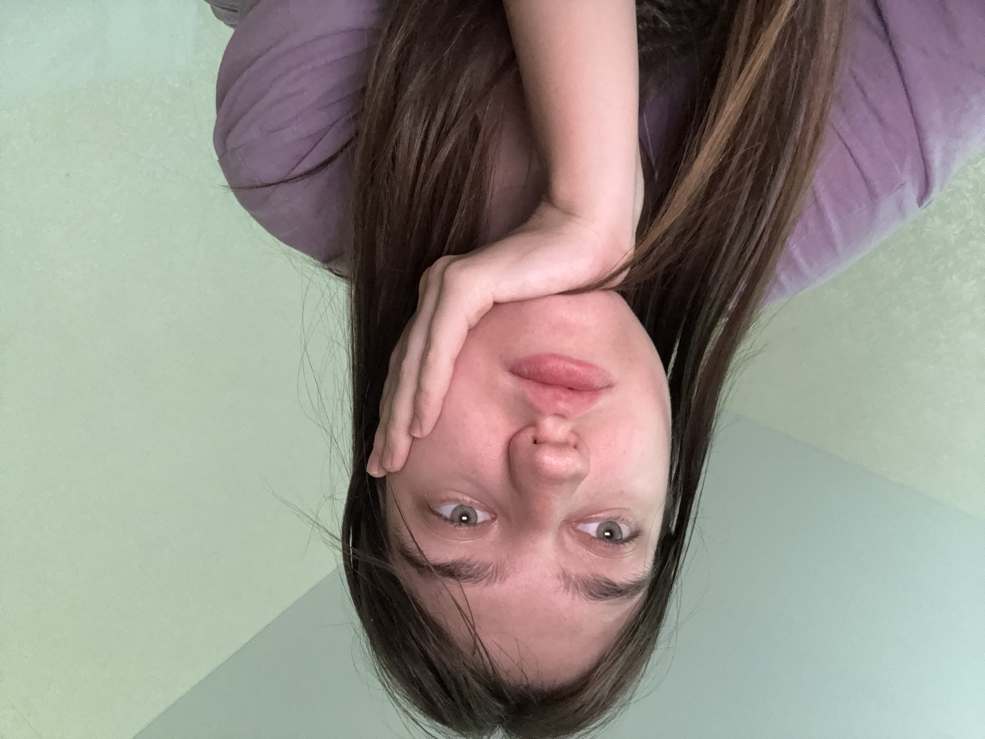 A woman with brown hair is pictured from a low, upside-down angle. She is wearing a purple shirt. Her right hand is resting on her face. The background is a two-tone wall, with a lighter shade on the right and a darker shade on the left. The location is Unknown, Unknown.
