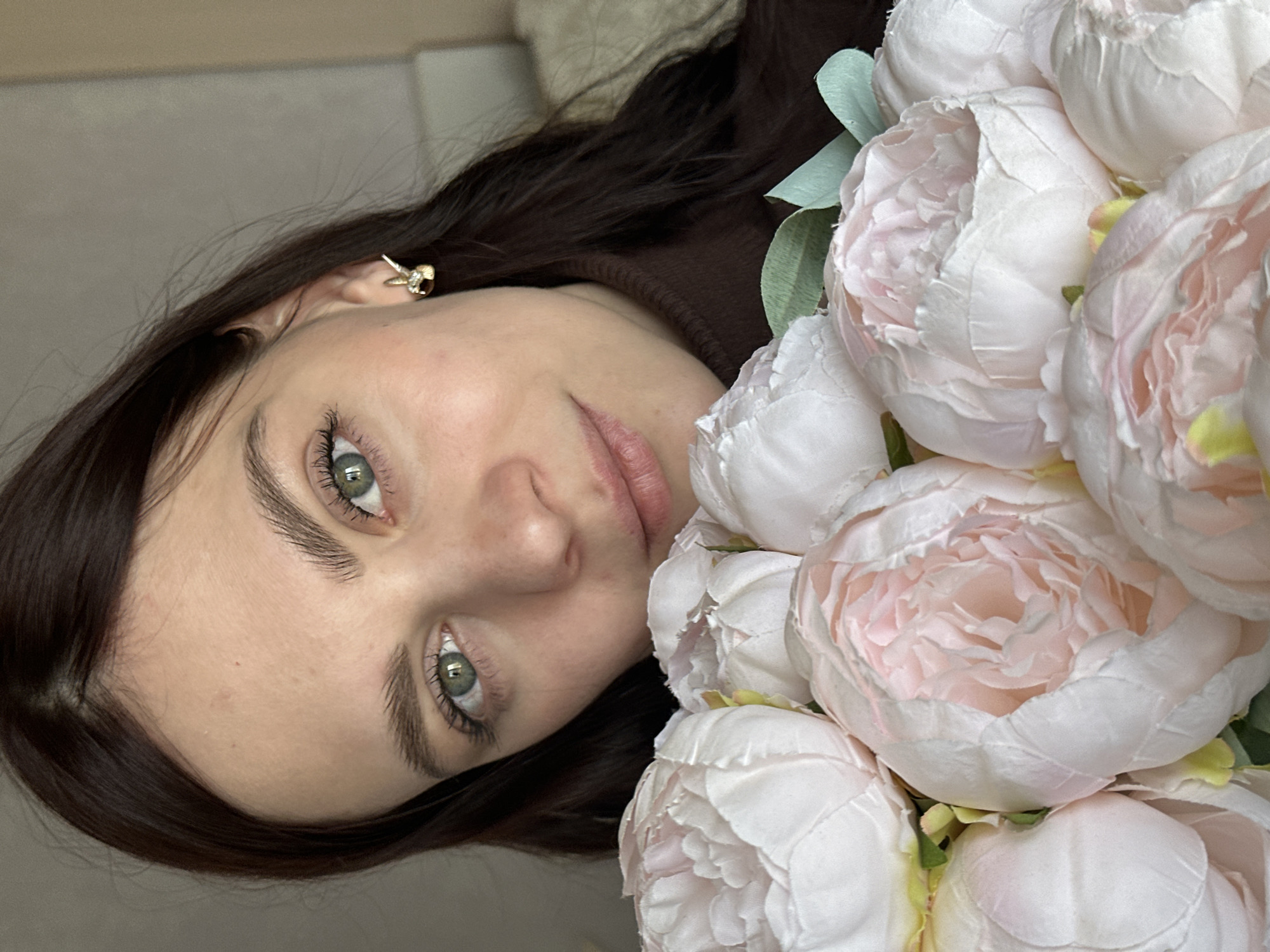 A woman with light skin, dark hair, and green eyes is positioned beside a bouquet of large, light pink and white peonies. She wears a brown turtleneck sweater and a gold earring in her left ear. The location is unknown and cannot be determined from the image.