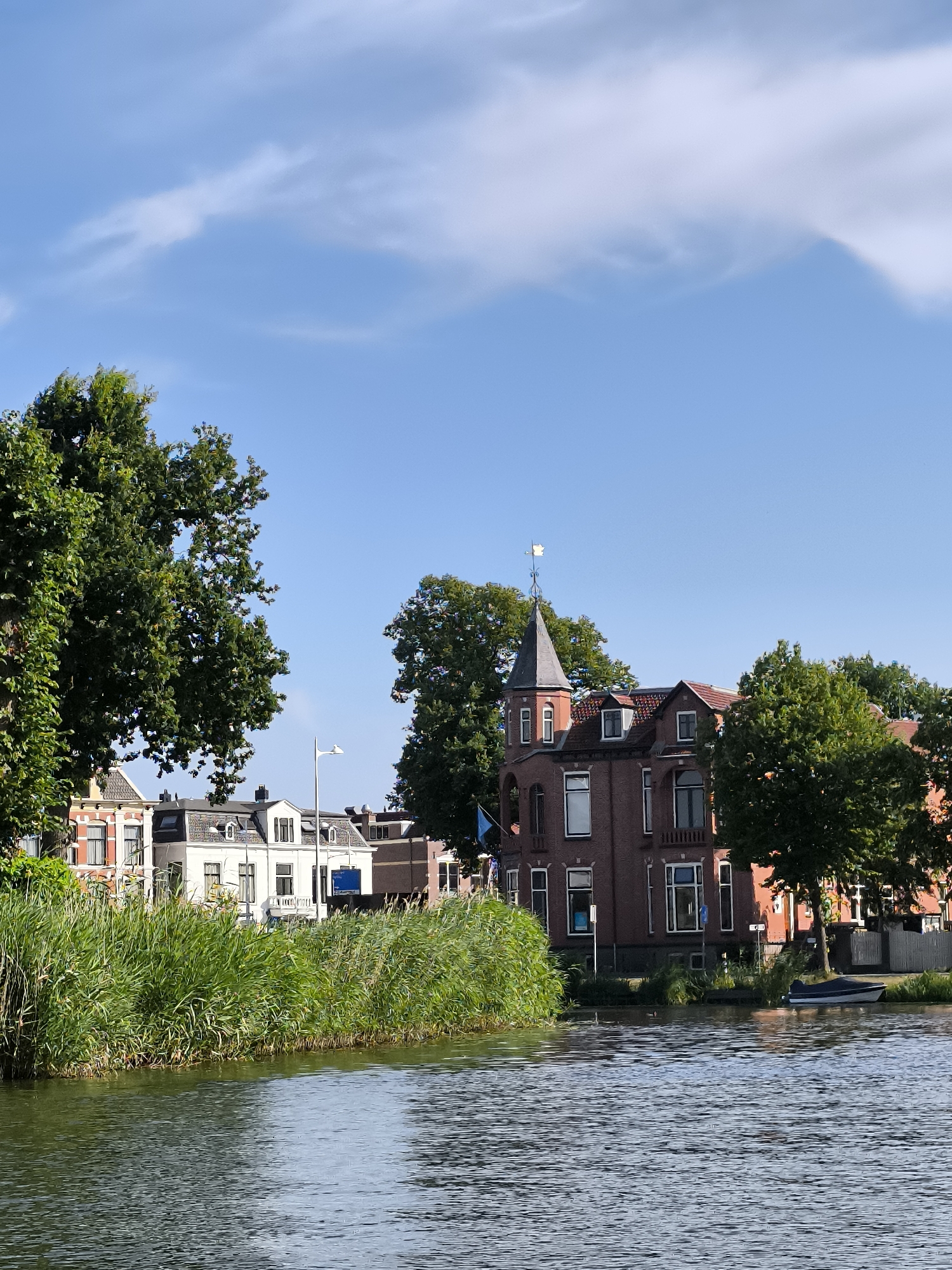 This is a daytime, eye-level, medium shot of a canal in Alkmaar, Netherlands, with buildings lining the far bank. In the foreground, the water is rippled, reflecting the sky. Tall, green reeds grow along the edge of the canal. Behind the reeds, a row of houses and trees are visible.
The most prominent building is a red brick house with a pointed tower and a weather vane. To its left, there is a white house with a dark mansard roof. Further to the left, a red-brick house with white trim is partially visible. The houses appear to be residences.
Several trees with lush green foliage are scattered throughout the scene, providing shade and framing the buildings. The sky is blue with scattered white clouds.
On the water, a small dark blue boat with a white cover is moored near the bank. A single duck is visible on the water between the reeds and the boat. There are no people visible in the image. The environment suggests a peaceful, urban waterway setting.