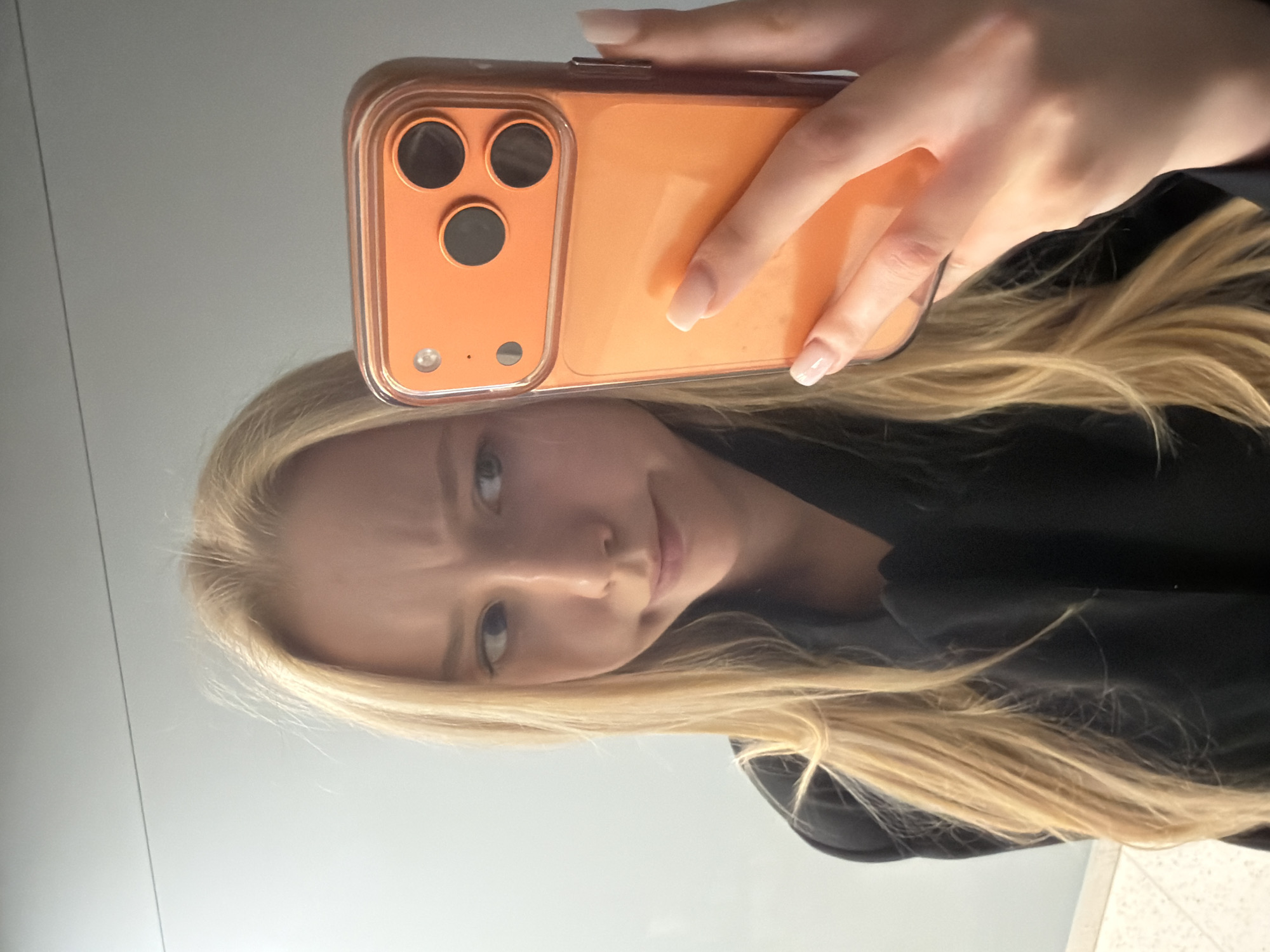A woman with long blonde hair and fair skin is holding an orange smartphone in front of her face, presumably taking a selfie in a mirror. The environment appears to be indoors with a plain, light-colored wall. She is wearing a black top. The location is unknown.