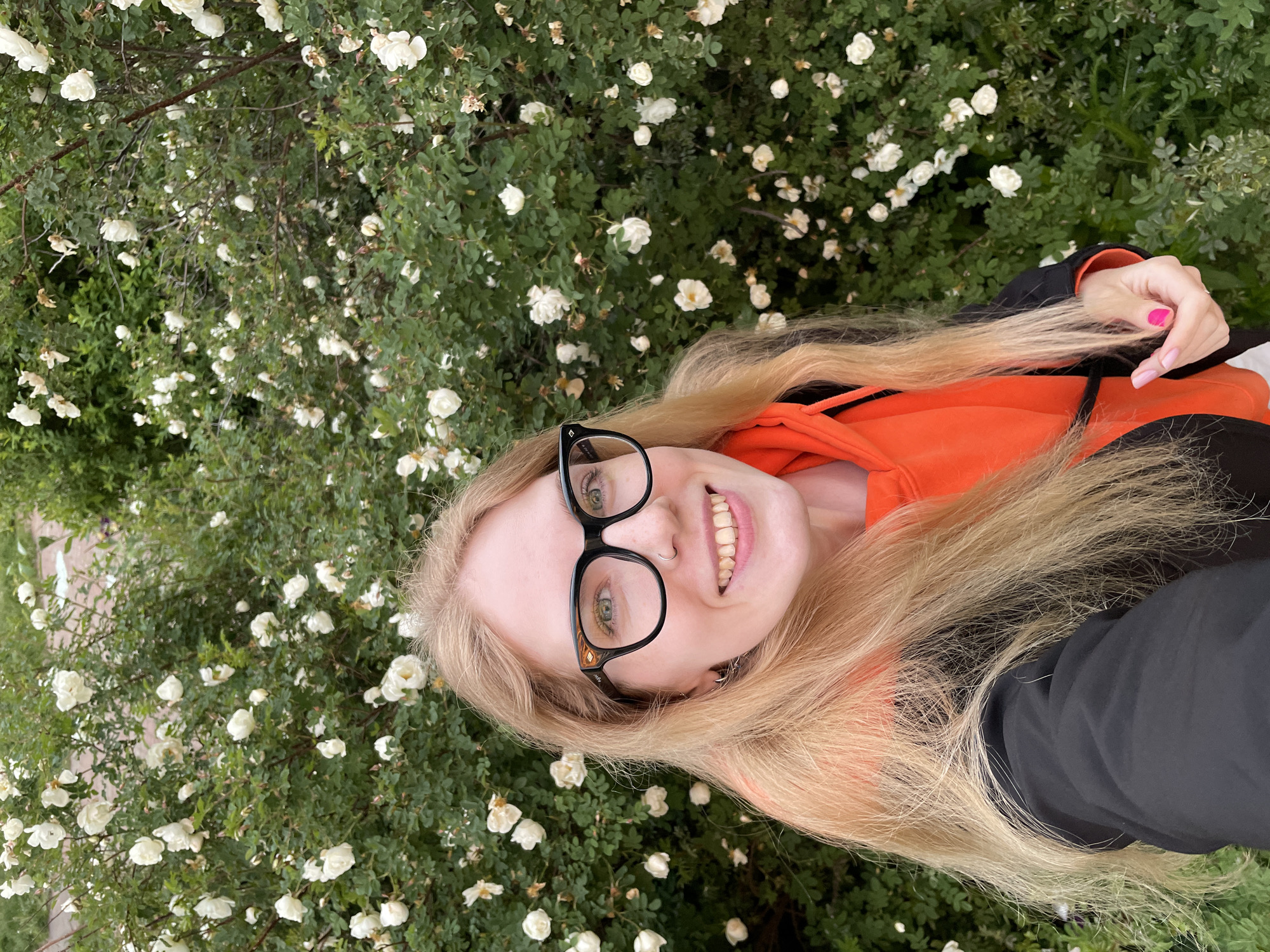 A young woman with long blonde hair and black-rimmed glasses is captured in a selfie, smiling broadly at the camera. She is wearing a bright orange hoodie under a black jacket. Her left hand, adorned with pink and light pink nail polish, rests on her hair near her shoulder. A small nose ring is visible on her right nostril.
The woman is positioned horizontally across the frame, suggesting she might be lying down or leaning back against something, with her head tilted upwards. The background is dominated by a dense, leafy green bush covered in numerous small, white flowers, resembling wild roses. The setting appears to be outdoors, likely in a park or garden, consistent with Izhevsk, Russia. The lighting is soft, indicating it is daytime, possibly overcast or late afternoon. No visible text is discernible in the image. The overall mood is cheerful and relaxed.