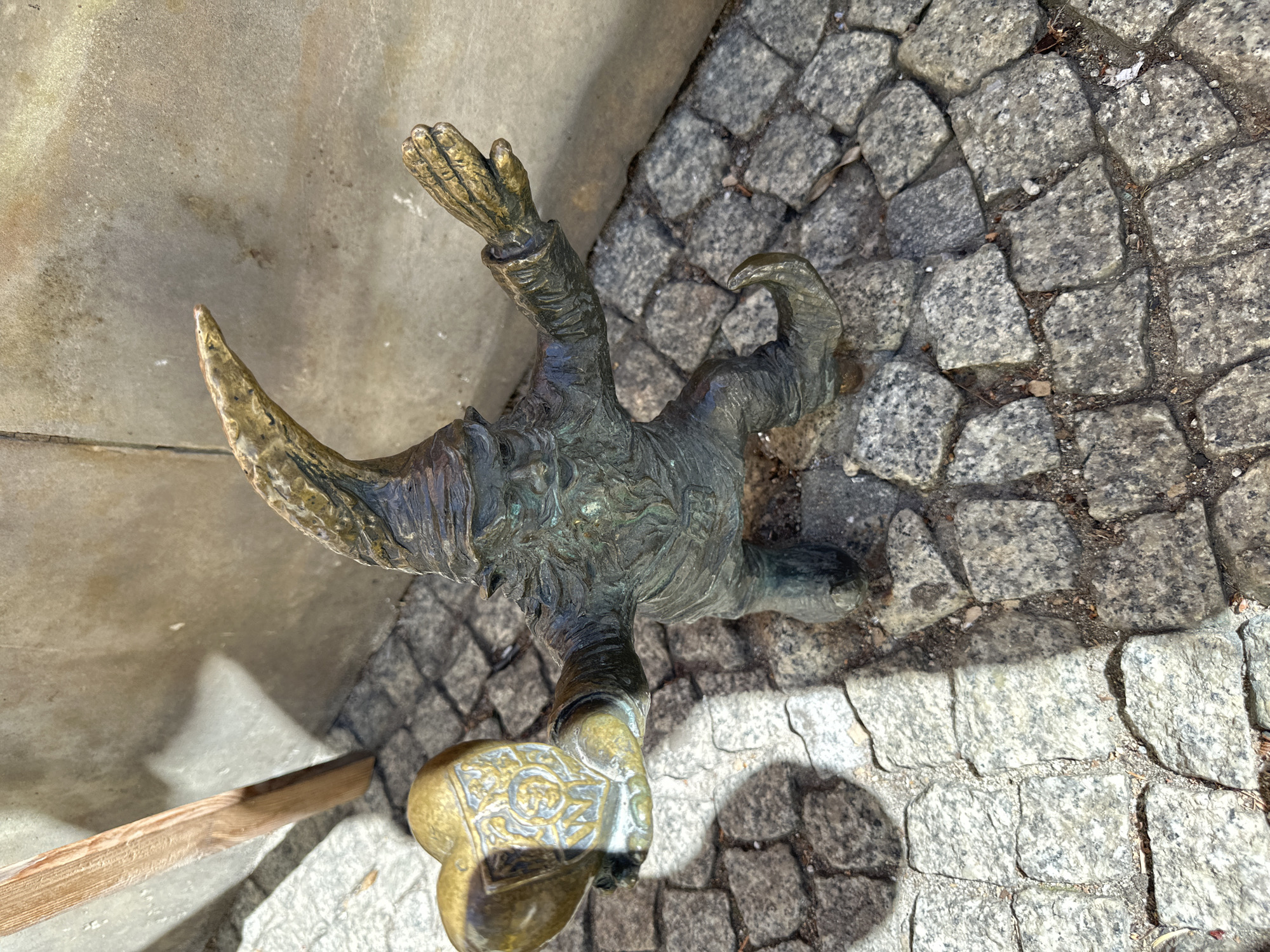A bronze statue of a Wrocław dwarf stands on a cobblestone pavement. The dwarf has a pointed hat and a smiling face, with one hand raised in greeting and the other hand resting on his belly. The cobblestones are gray and vary in size. A stone or concrete surface is to the left of the figure. The light suggests it's a sunny day. The location is likely Wrocław, Poland, where these dwarf statues are a popular tourist attraction.