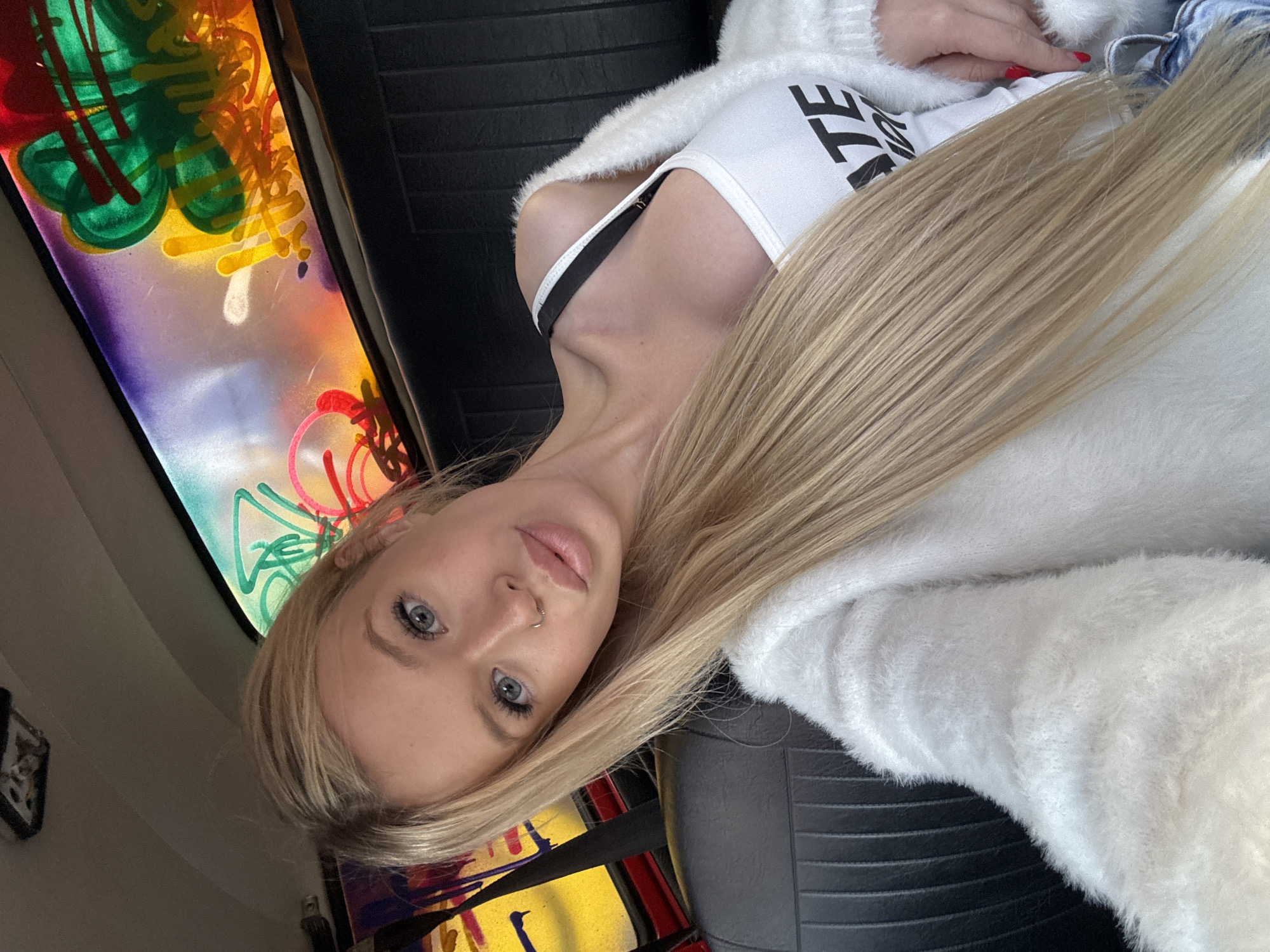 A young blonde woman with blue eyes and a nose ring takes a selfie inside a car. She is wearing a white tank top with black text that reads "ATE" and a fluffy white cardigan. Her long blonde hair is falling over her right shoulder. The car's interior is visible, including a black seat and a ceiling light. The background features a colorful graffiti mural on what appears to be the car's rear window or partition. The lighting suggests it is daytime. The location is specified as Saint Petersburg, Russia.